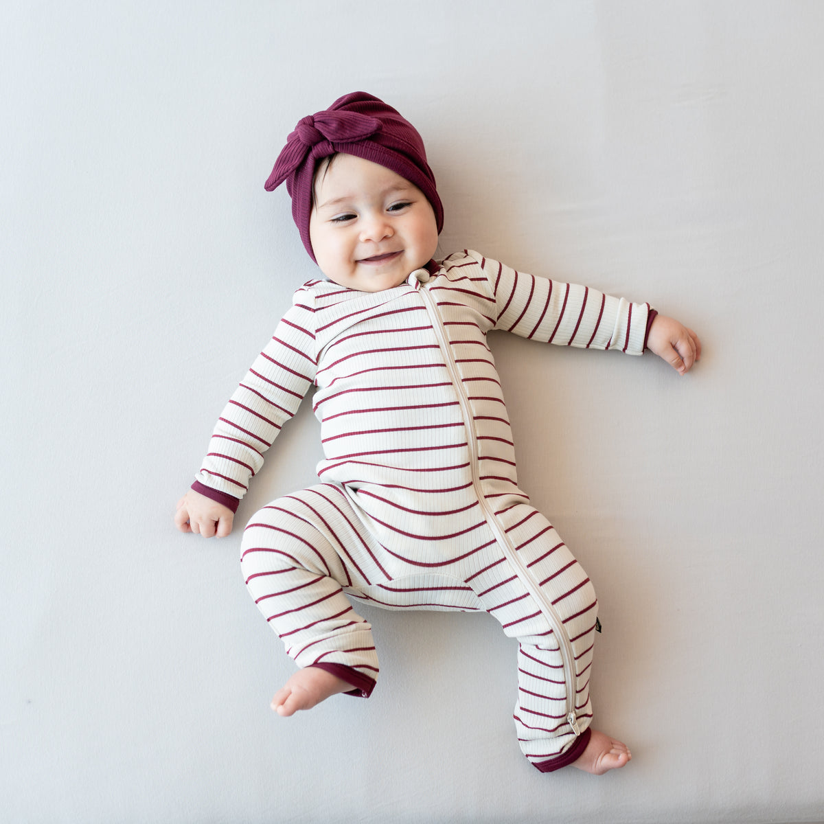 Smiling infant laying down wearing the Ribbed Zipper Romper in Burgundy Stripe and the Ribbed Headwrap in Burgundy