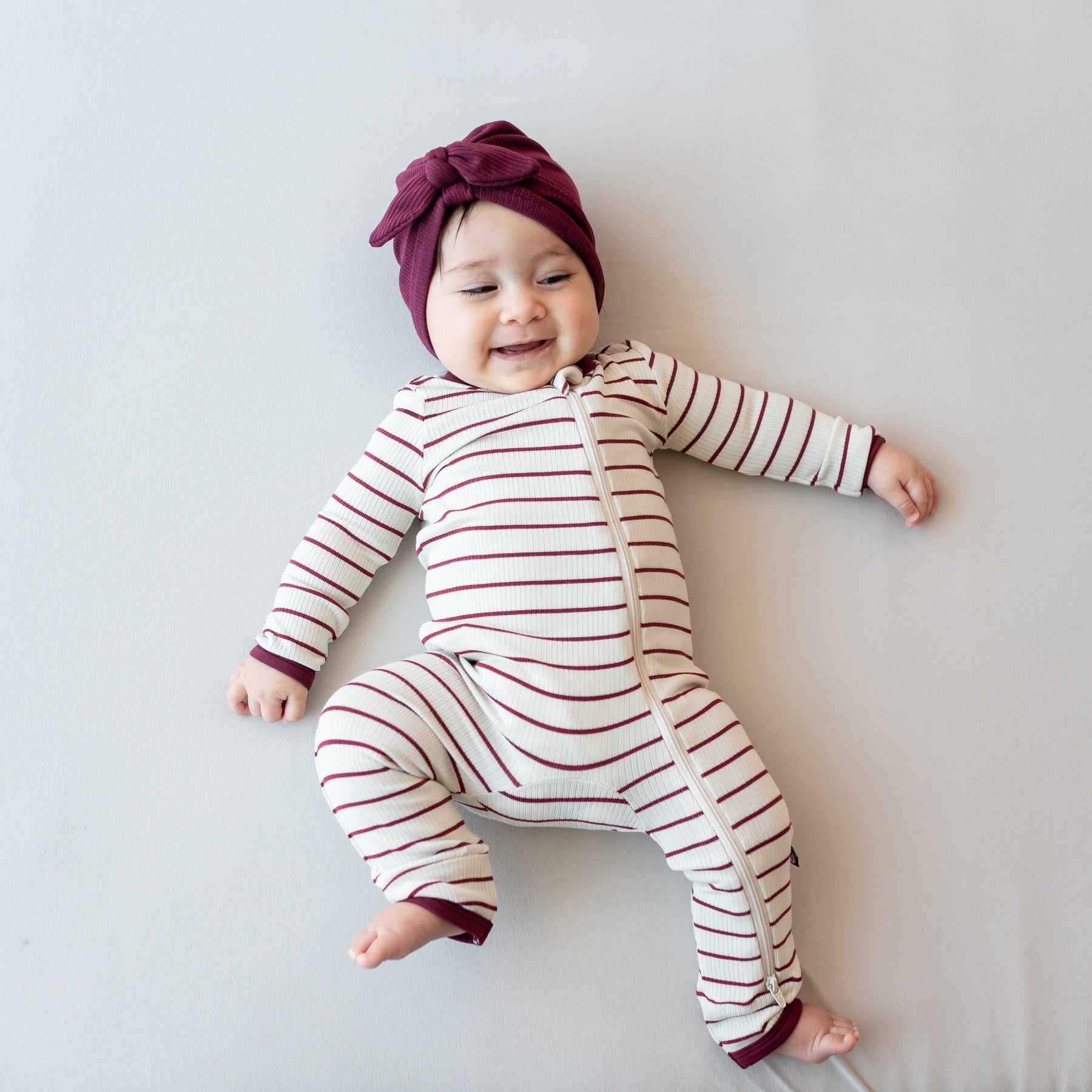 Smiling infant laying on a light neutral background wearing the Ribbed Zipper Romper in Burgundy Stripe and ribbed headwrap in burgundy