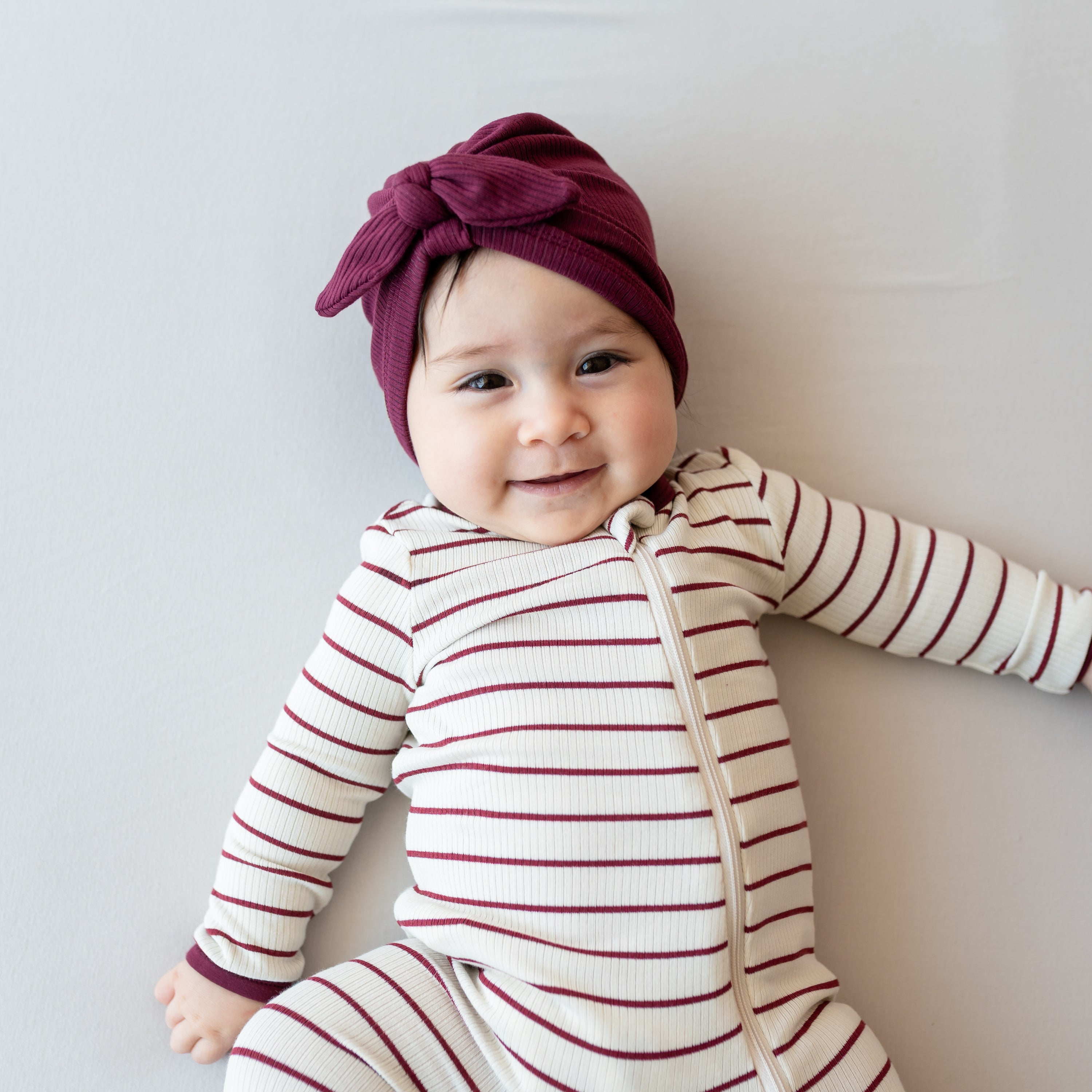 Close up of a smiling infant laying down wearing the Ribbed Zipper Romper in Burgundy Stripe and Burgundy ribbed headwrap