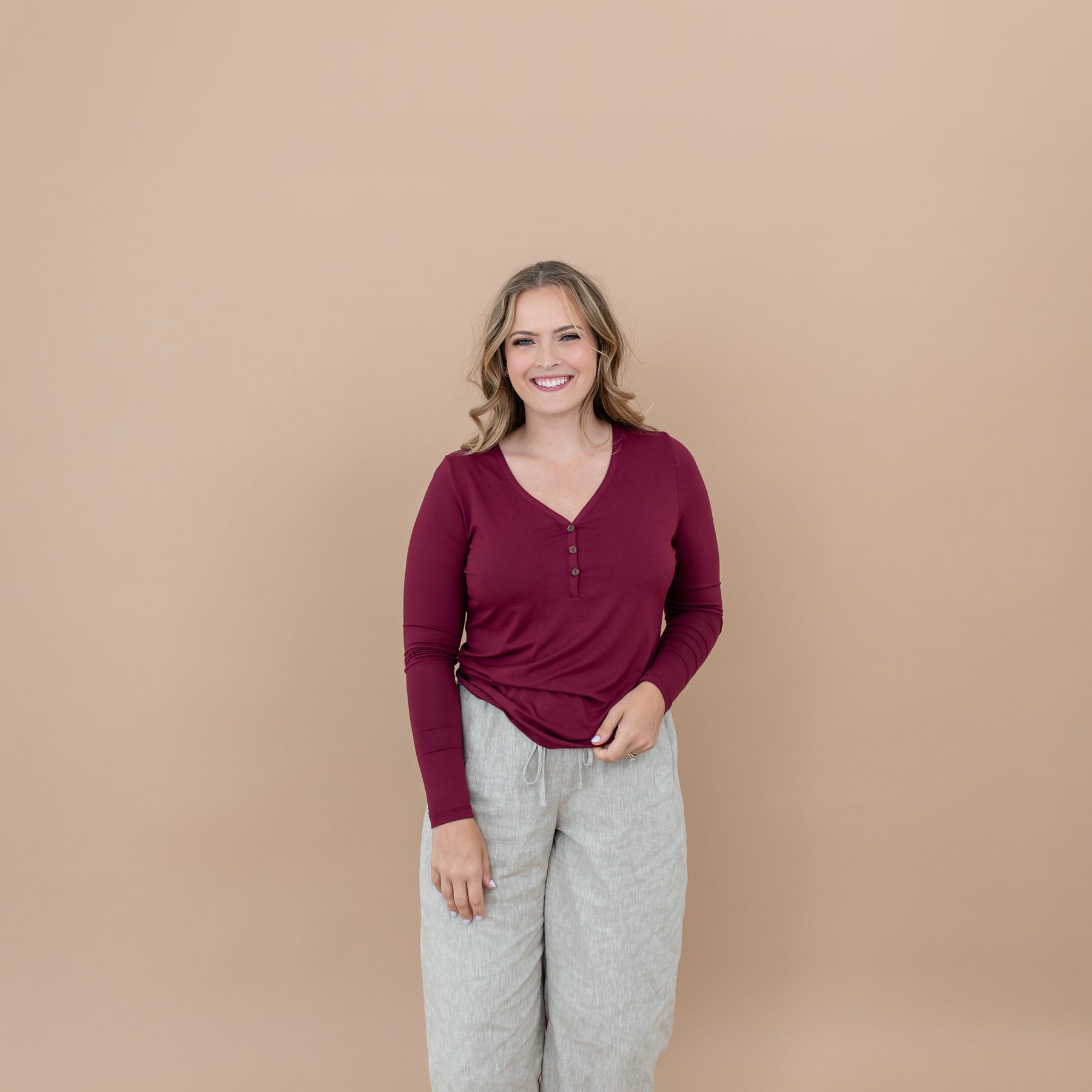 Smiling female model wearing the Women's Long Sleeve Henley Tee in Burgundy paired with a linen pant