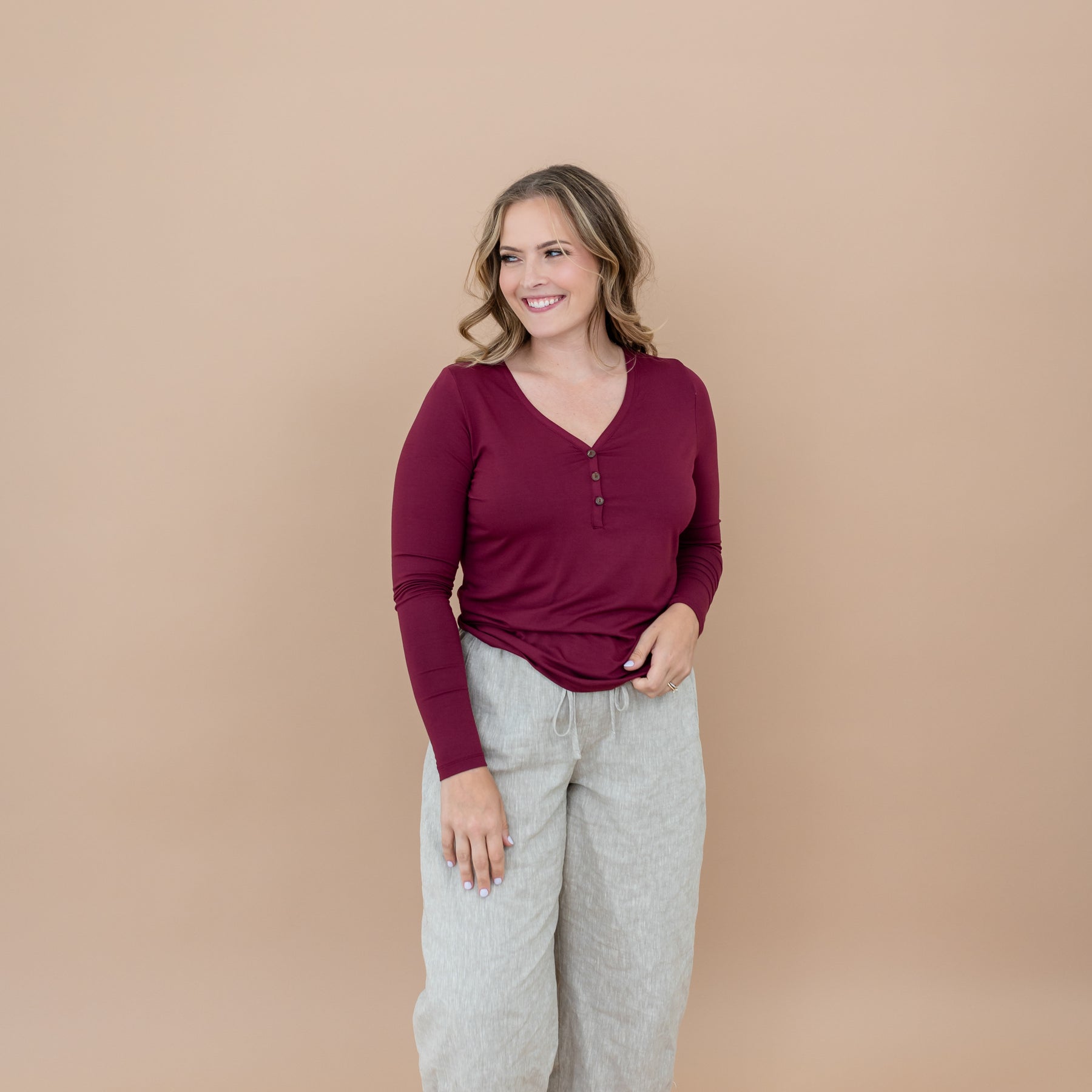Smiling female model looking off to the side wearing the Women's Long Sleeve Henley Tee in Burgundy paired with a linen pant