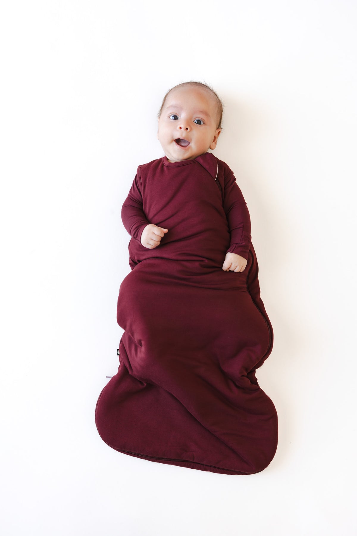 A baby lying on their back, wearing a deep burgundy wearable 1.0 TOG blanket, against a plain white background.