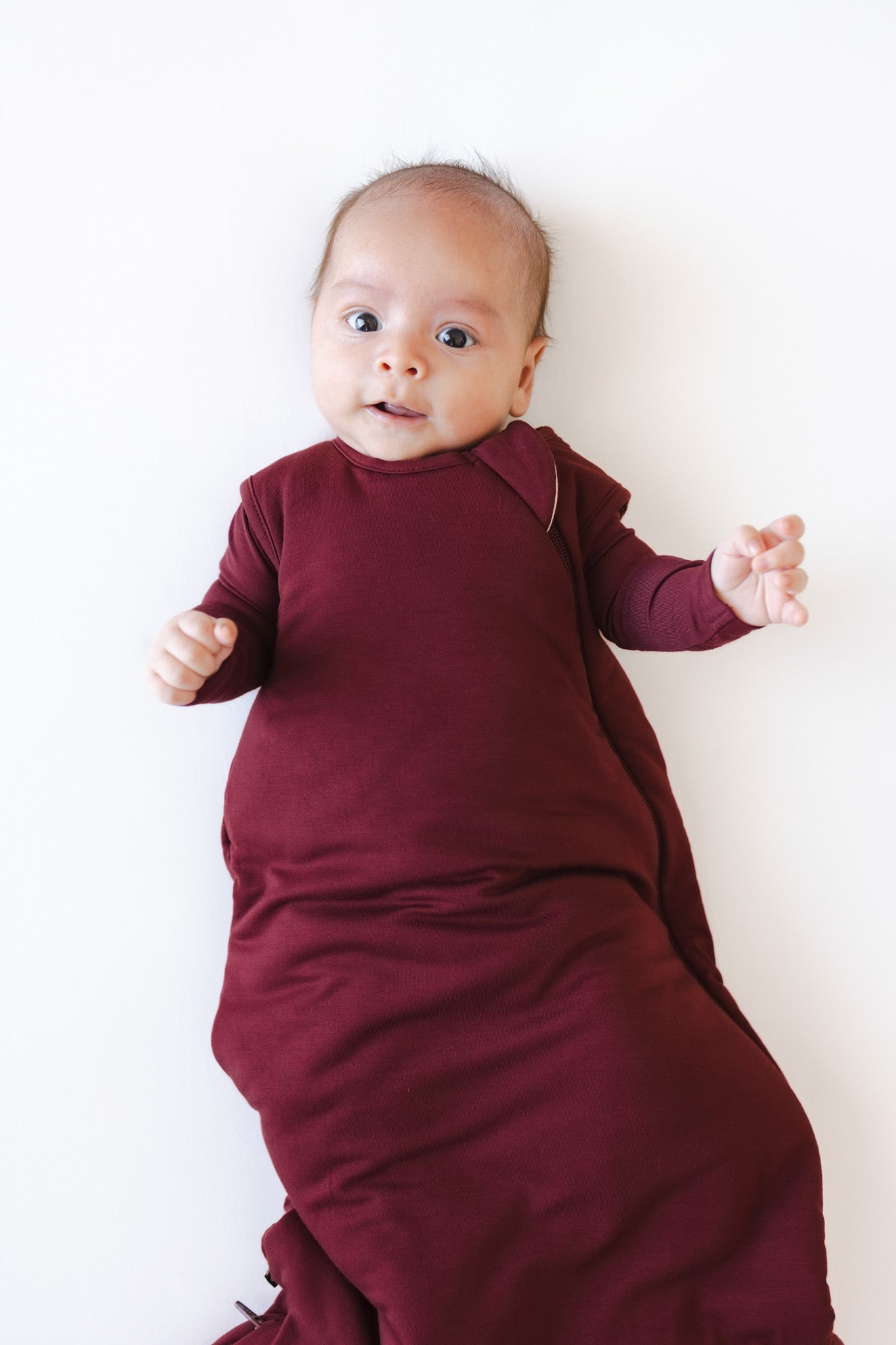 A baby lying on their back wearing a burgundy sleep sack against a plain white background.