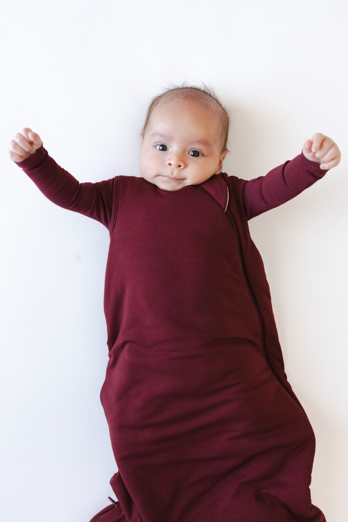 A baby lying on their back wearing a burgundy sleep sack against a plain white background.