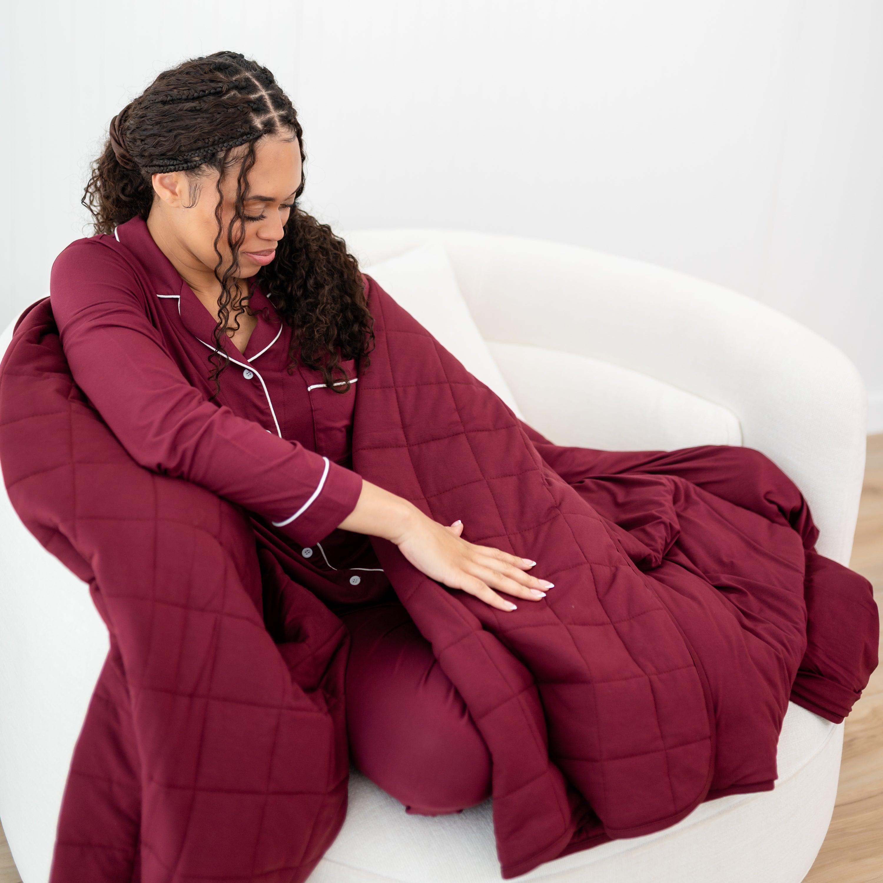 Female model sitting on a couch wrapped in the Adult Quilted Blanket in Burgundy 2.5 wearing the long sleeve pajama set in the matching color