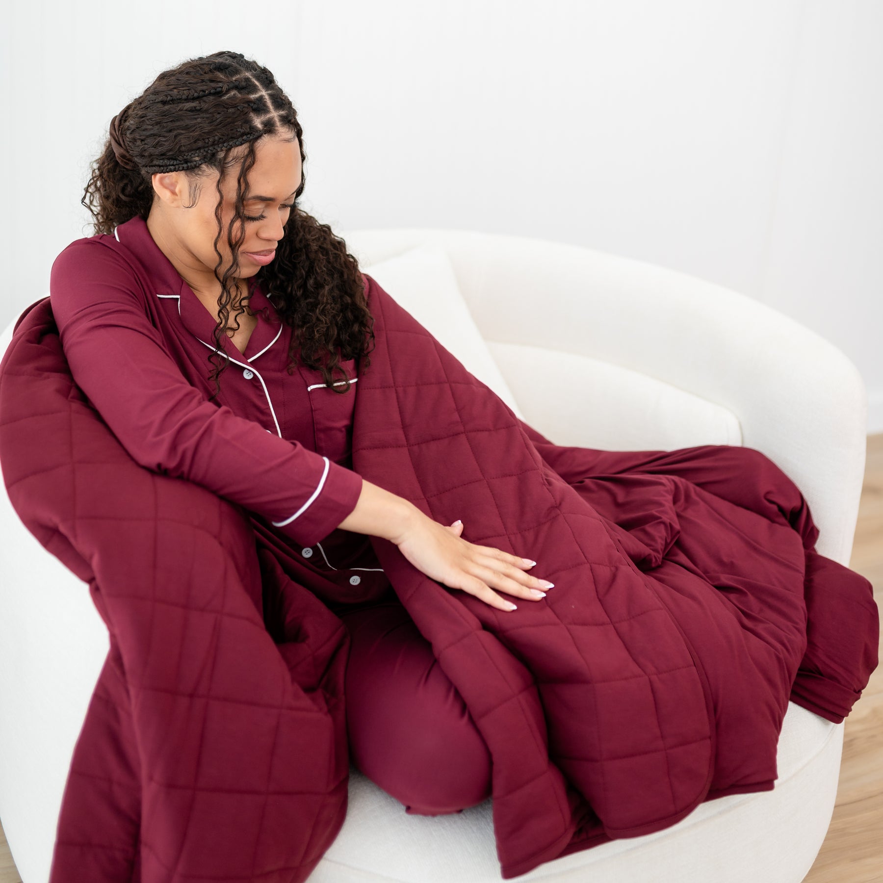 Female model sitting on a couch wrapped in the Adult Quilted Blanket in Burgundy 2.5 wearing the long sleeve pajama set in the matching color