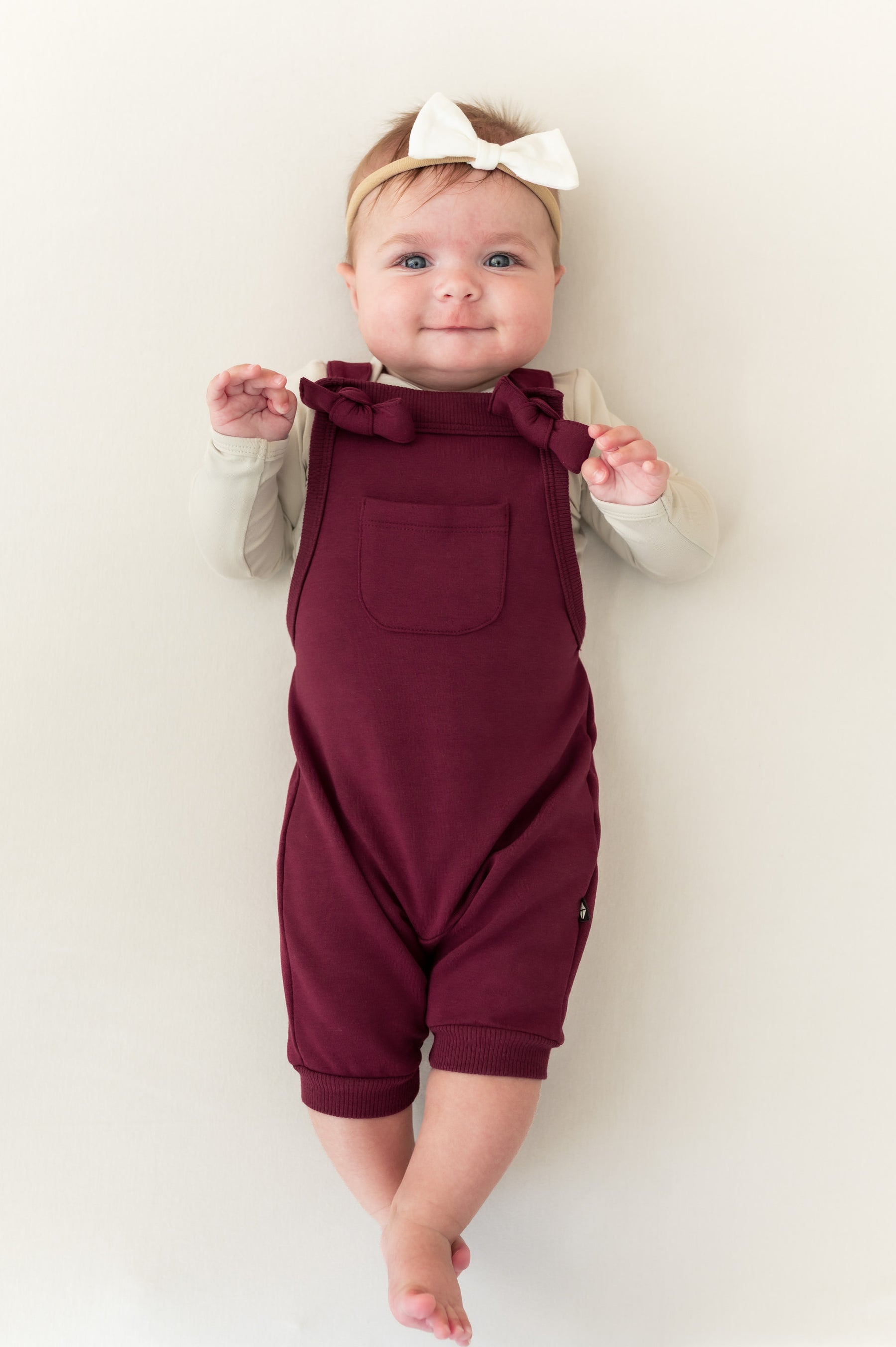 Baby wearing Overall in Burgundy color with white bow headband lying on a beige background