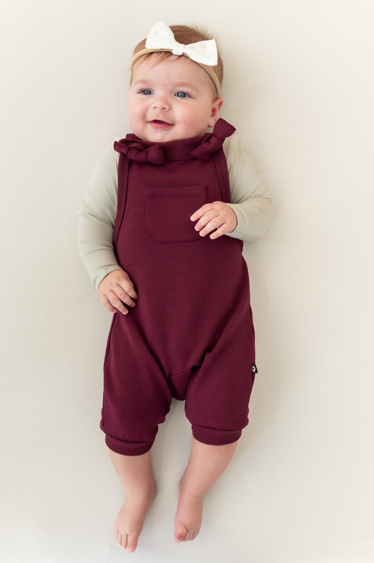 Baby wearing Overall in Burgundy color with white bow headband lying on a beige background