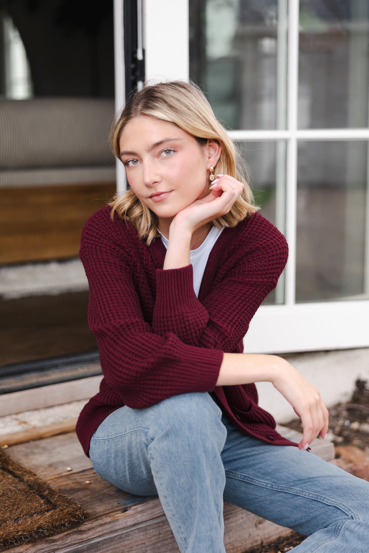 Modal sitting outdoors wearing Chunky Knit Women's Waffle Cardigan in Burgundy over white T-shirt and blue jeans