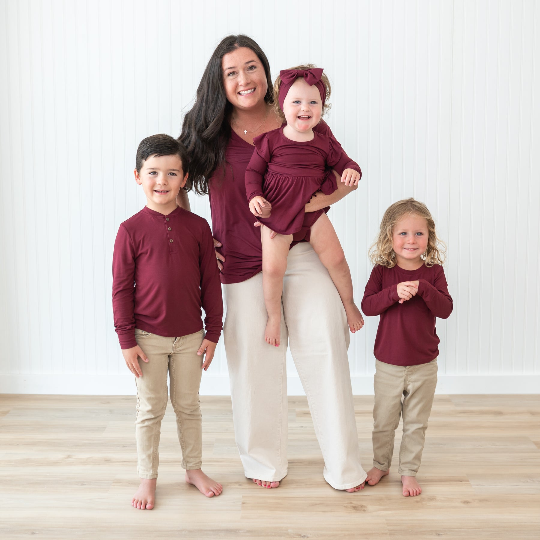 Family matching in Burgundy. Mother standing beside her two oldest children while holding her youngest. Pictures are the women's v-neck, toddler long sleeve henley, toddler long sleeve crew neck and twirl bodysuit dress