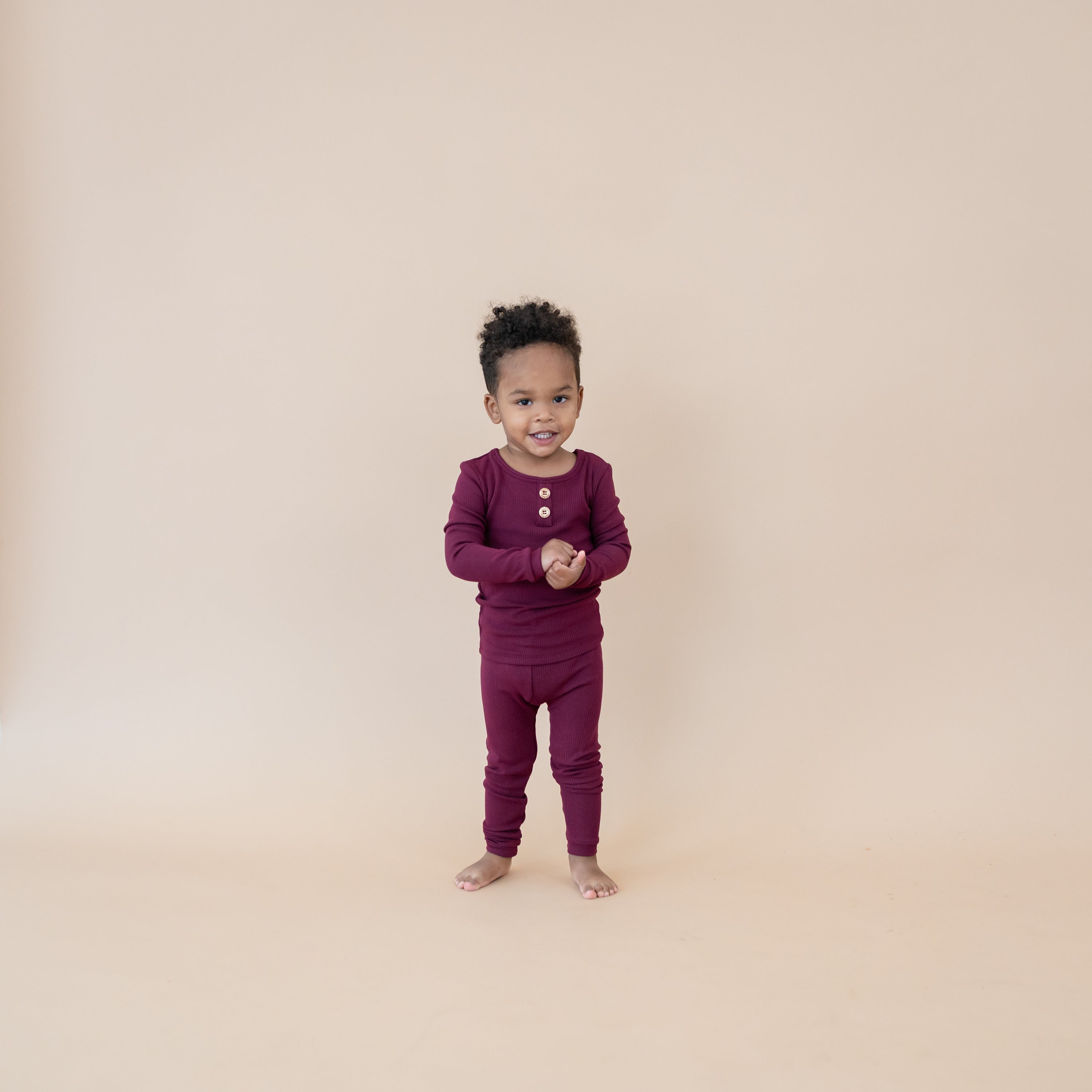 Toddler standing with hands together wearing the Ribbed Henley Set in Burgundy in front of a light taupe background