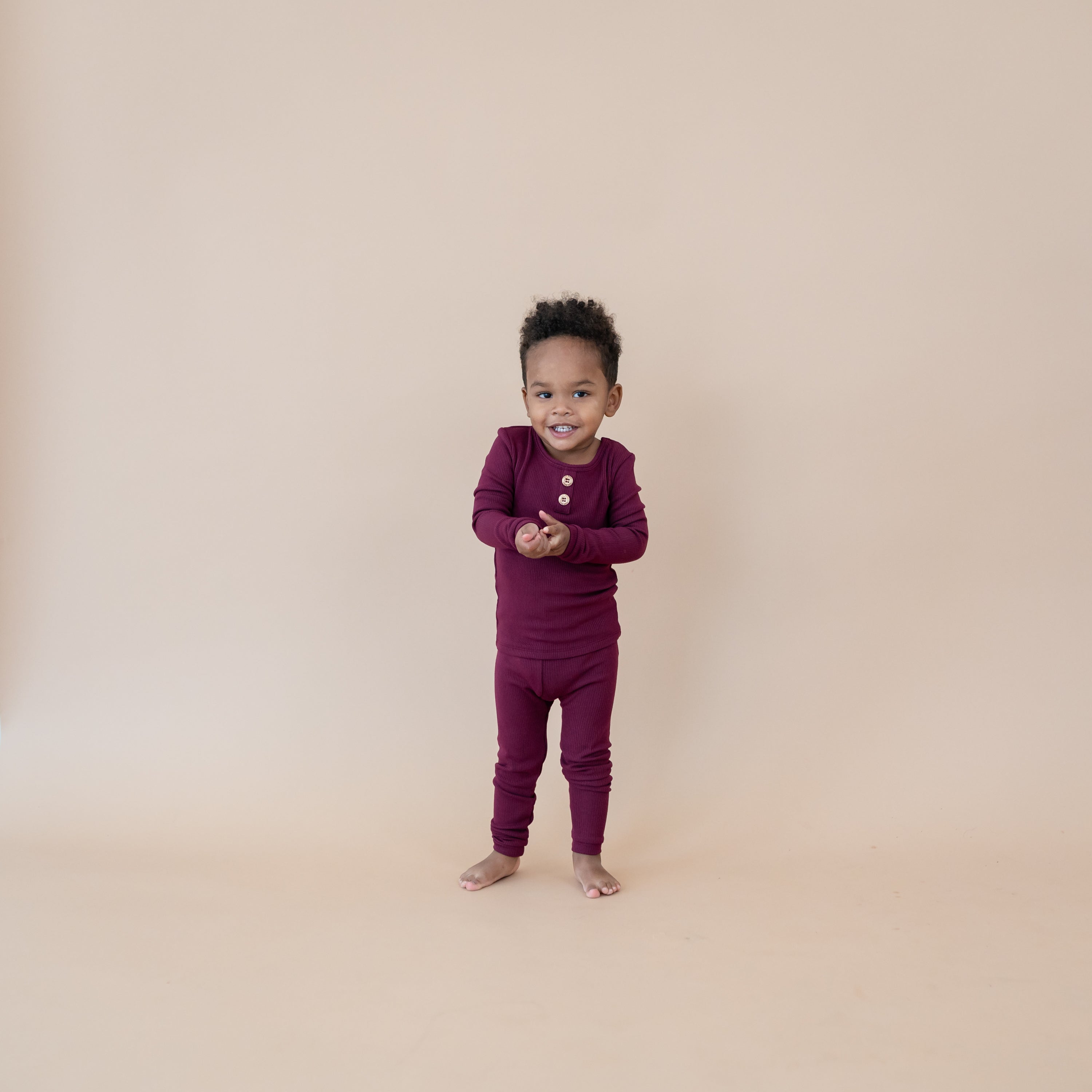 Toddler standing holding his hands in front of him wearing the Ribbed Henley Set in Burgundy with a taupe colored backdrop behind him