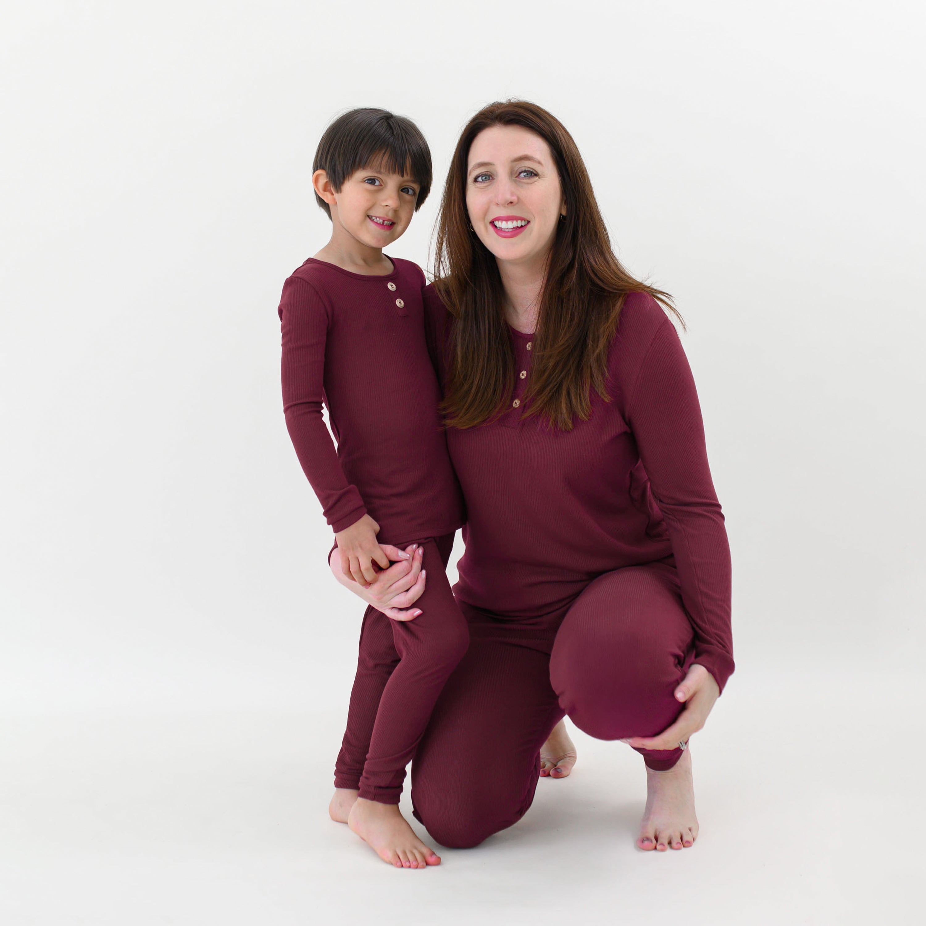 Mother kneeling beside her son with her arm around him wearing the Women’s Ribbed Henley Set in Burgundy in front of a white backdrop