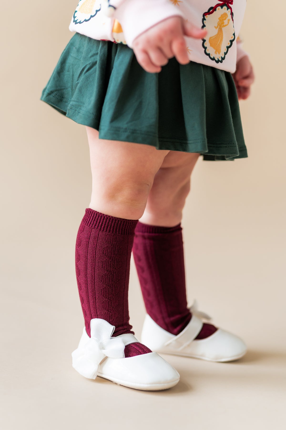 Toddler wearing Knee High Socks in Burgundy on white shoes under green skirt on a beige background