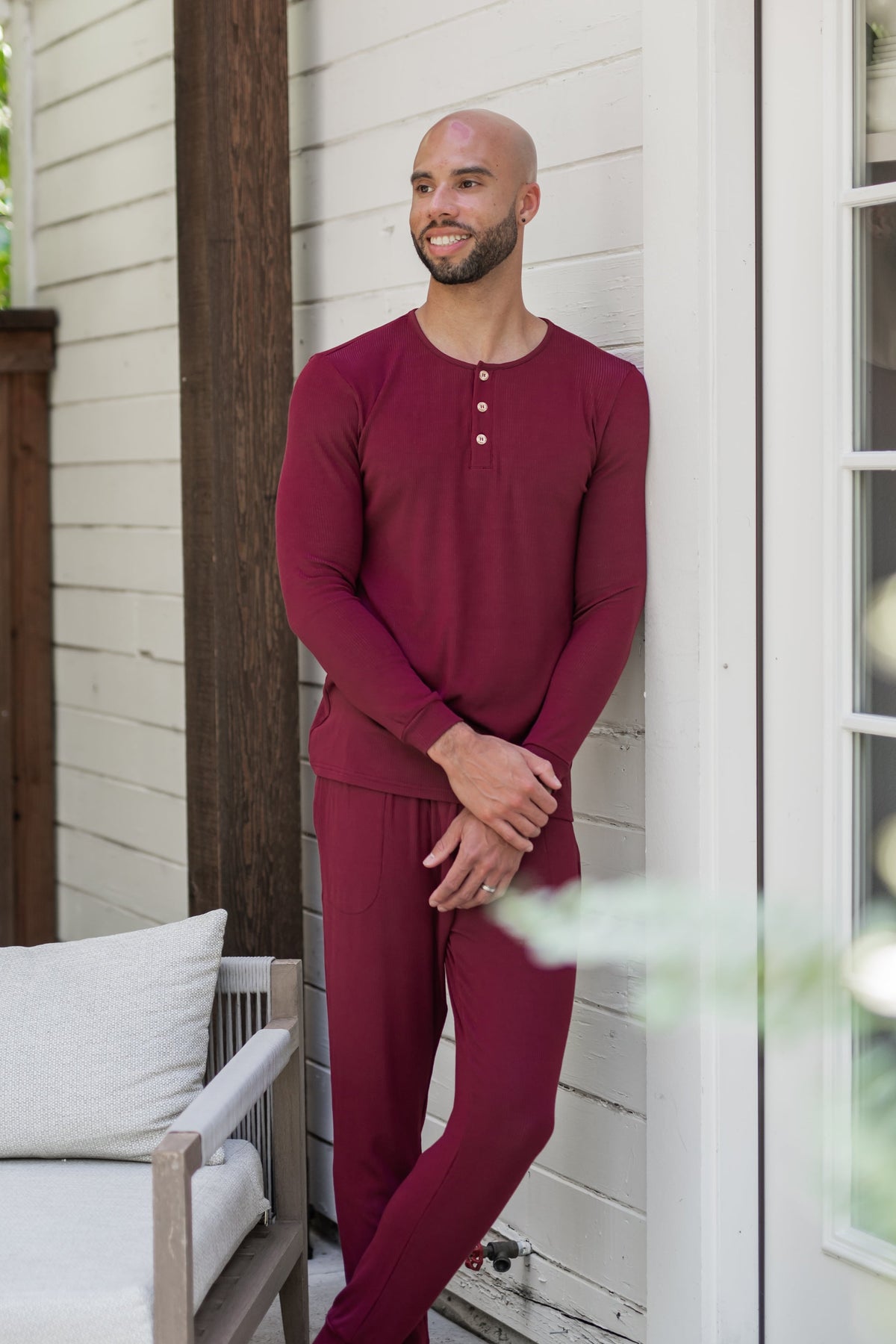 Male model leaning against a white house looking off to the side wearing the Men's Ribbed Henley Set in Burgundy