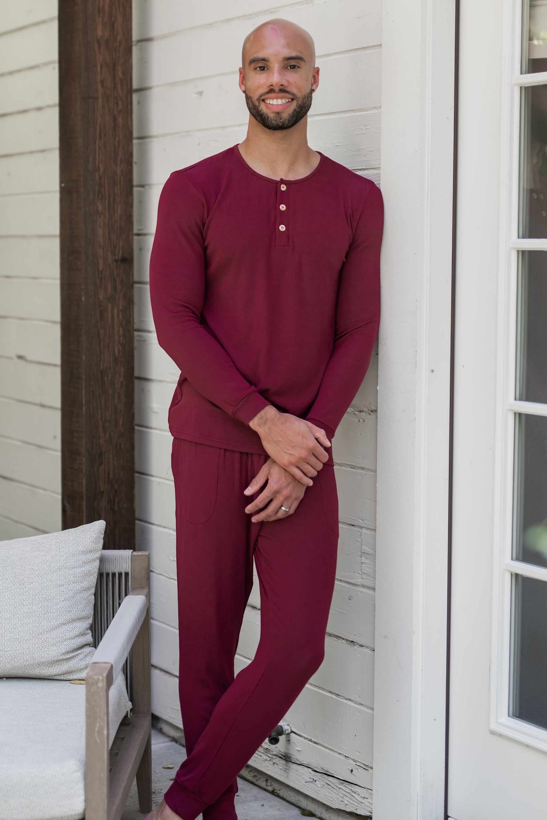 Male model leaning against a white house wearing the Men's Ribbed Henley Set in Burgundy