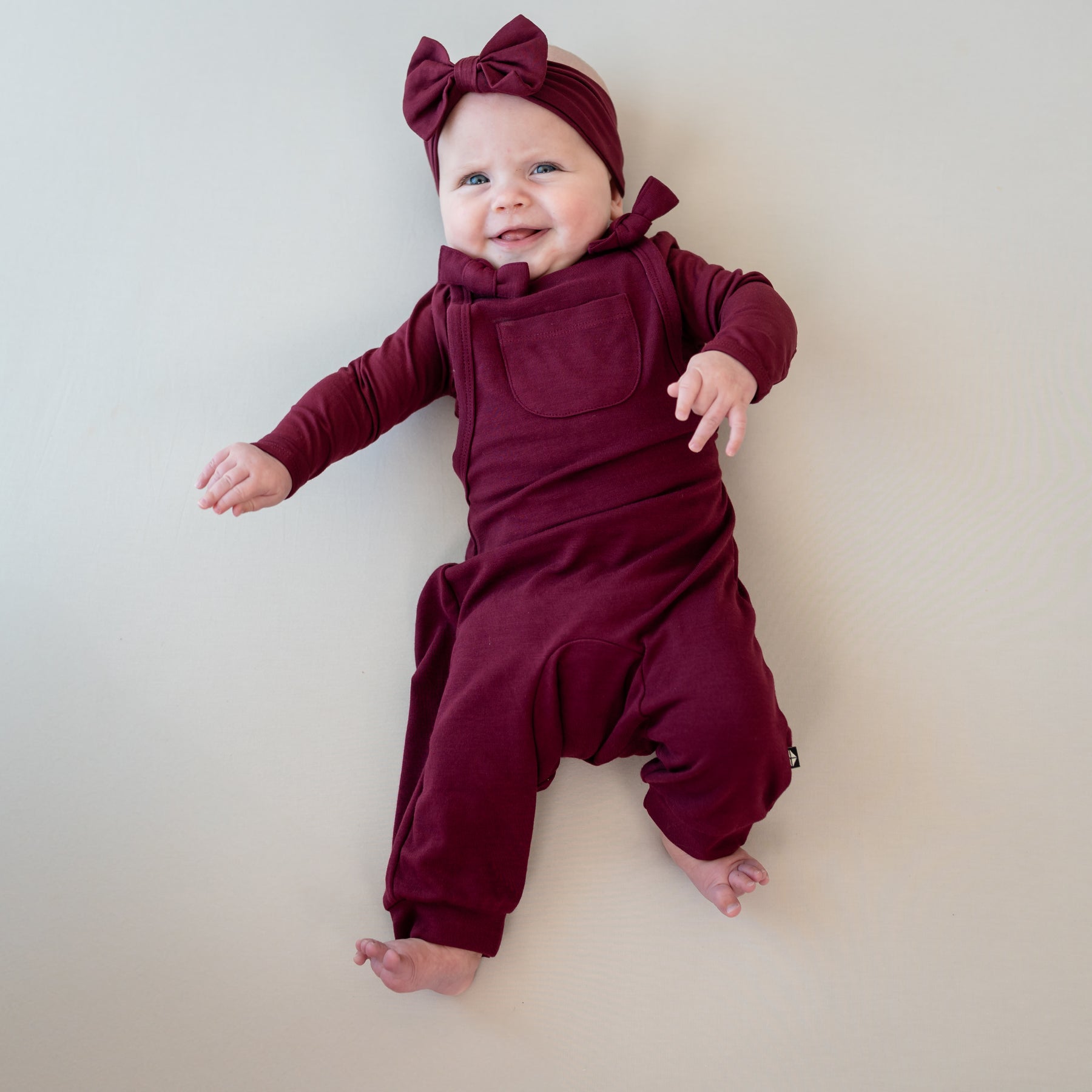 Smiling infant girl laying on a light neutral background with her hands in the air wearing the Bamboo Jersey Overall in Burgundy with matching long sleeve bodysuit and bow headband