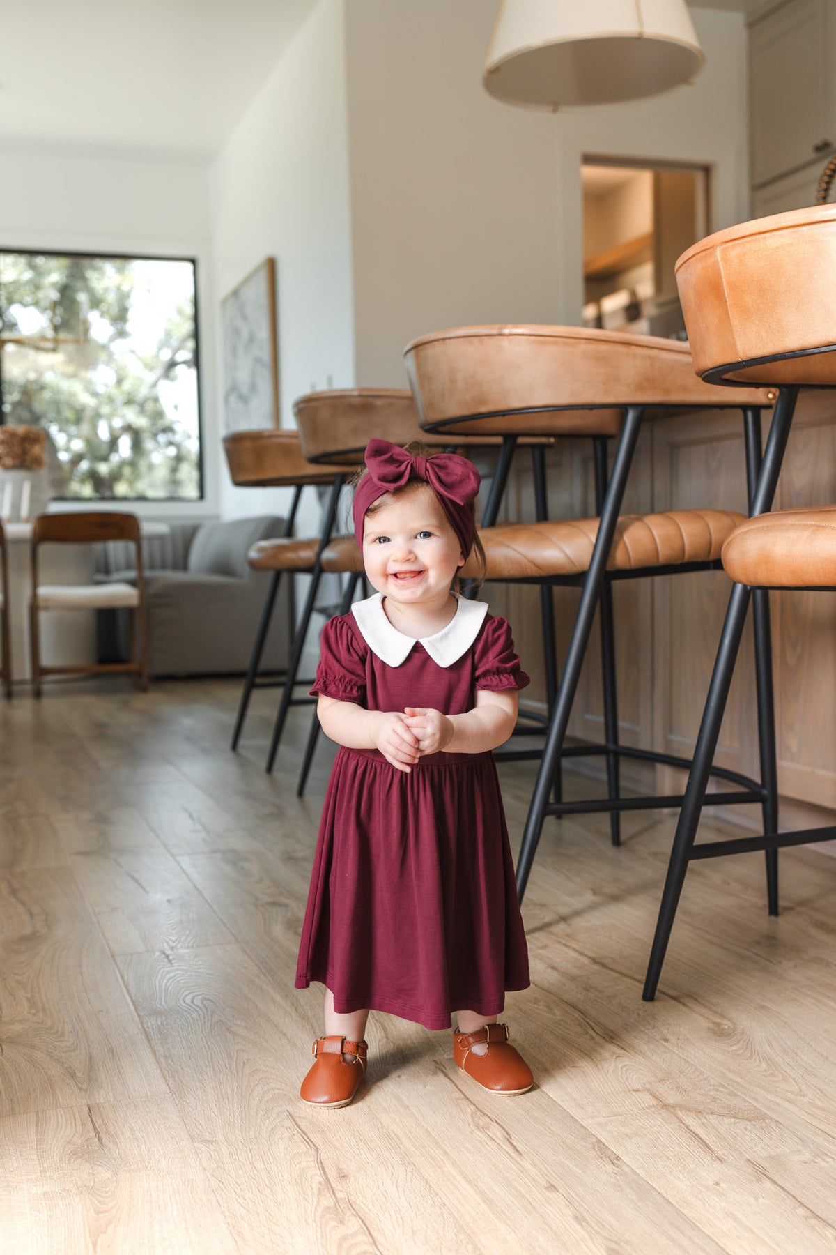 A toddler girl stands on a light wood floor in a modern kitchen, wearing a burgundy short-sleeve dress with a white Peter Pan collar, a matching burgundy bow headband, and brown leather shoes.