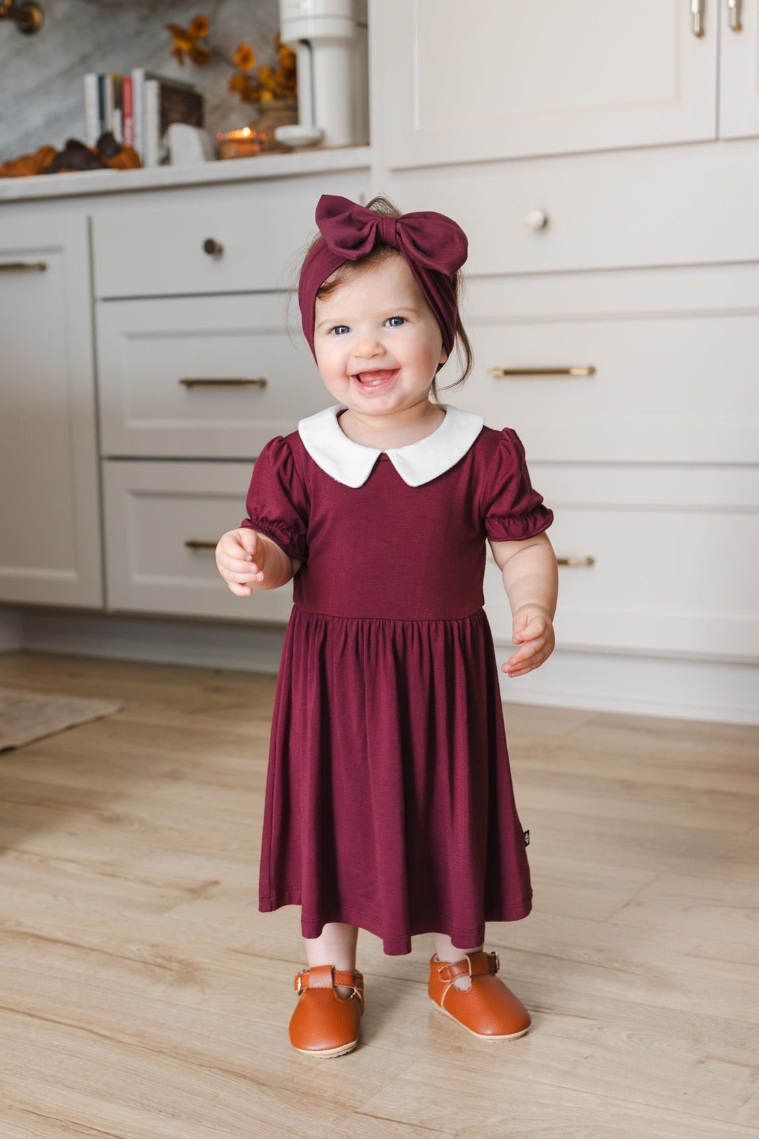 A toddler girl stands in a bright kitchen wearing a burgundy dress with a white Peter Pan collar, a matching bow headband, and tan shoes.