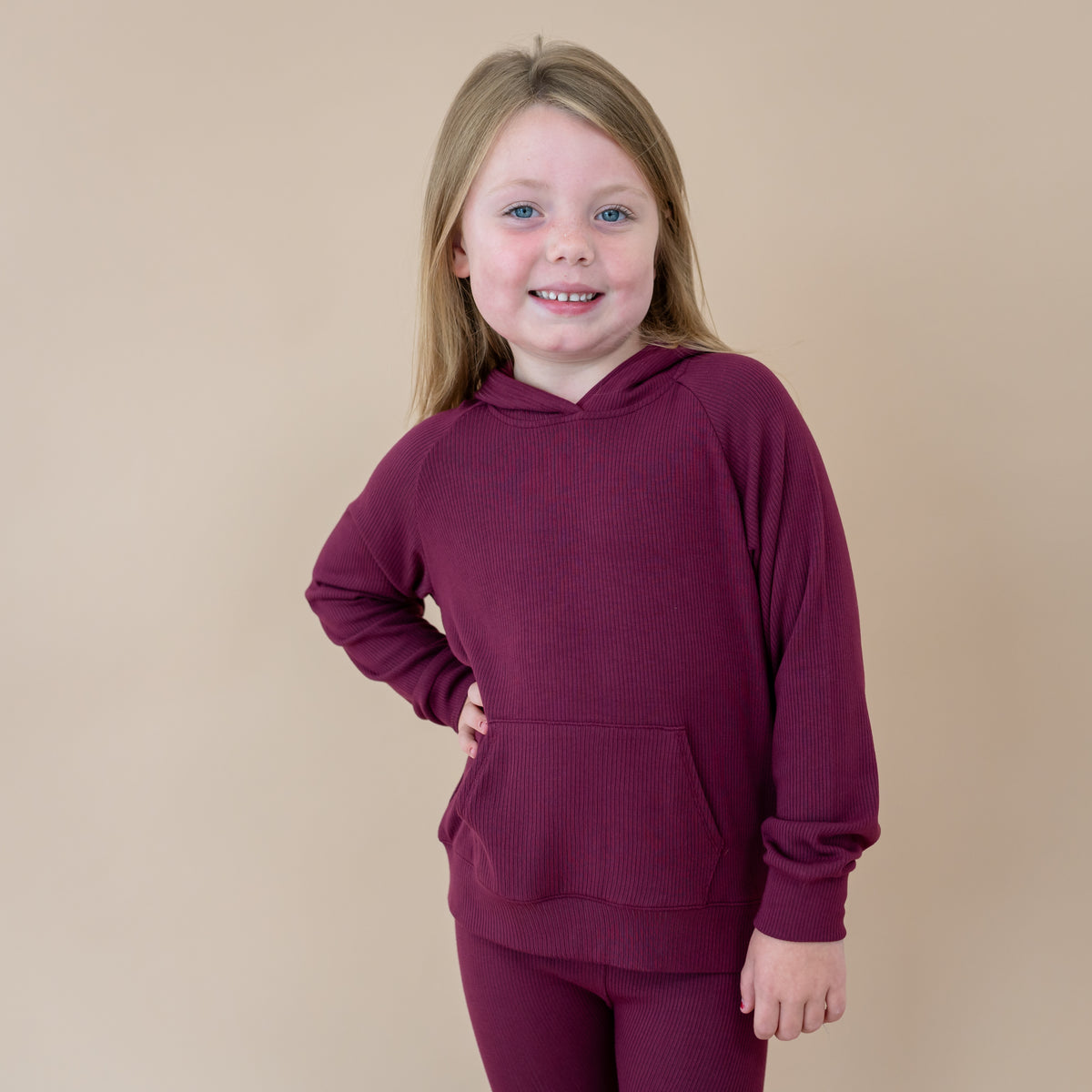 Young girl standing with her hand on her hip wearing the Ribbed Hoodie in Burgundy in front of a taupe backdrop