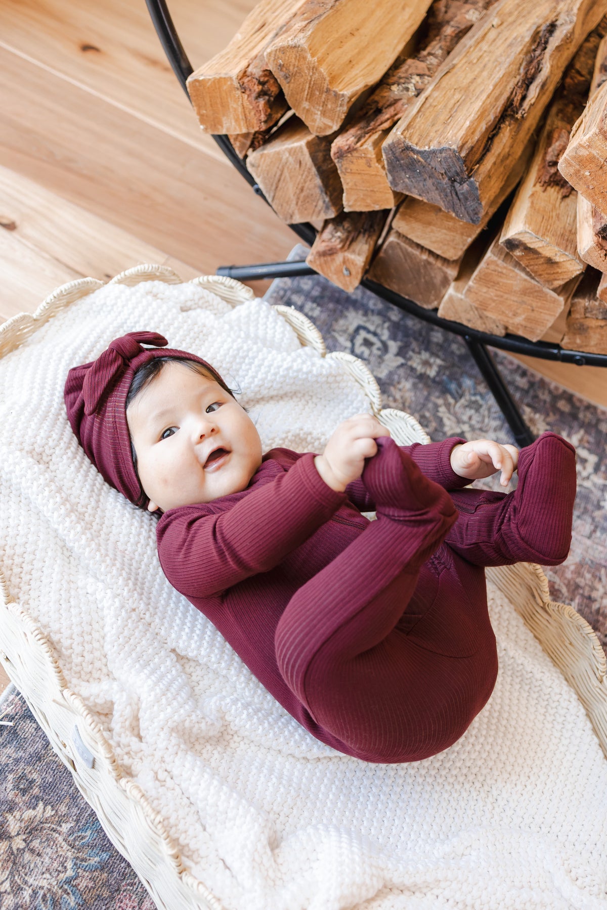 Baby holding her feet wearing Ribbed Zipper Footie and Ribbed Headwrap in Burgundy lying in a basket