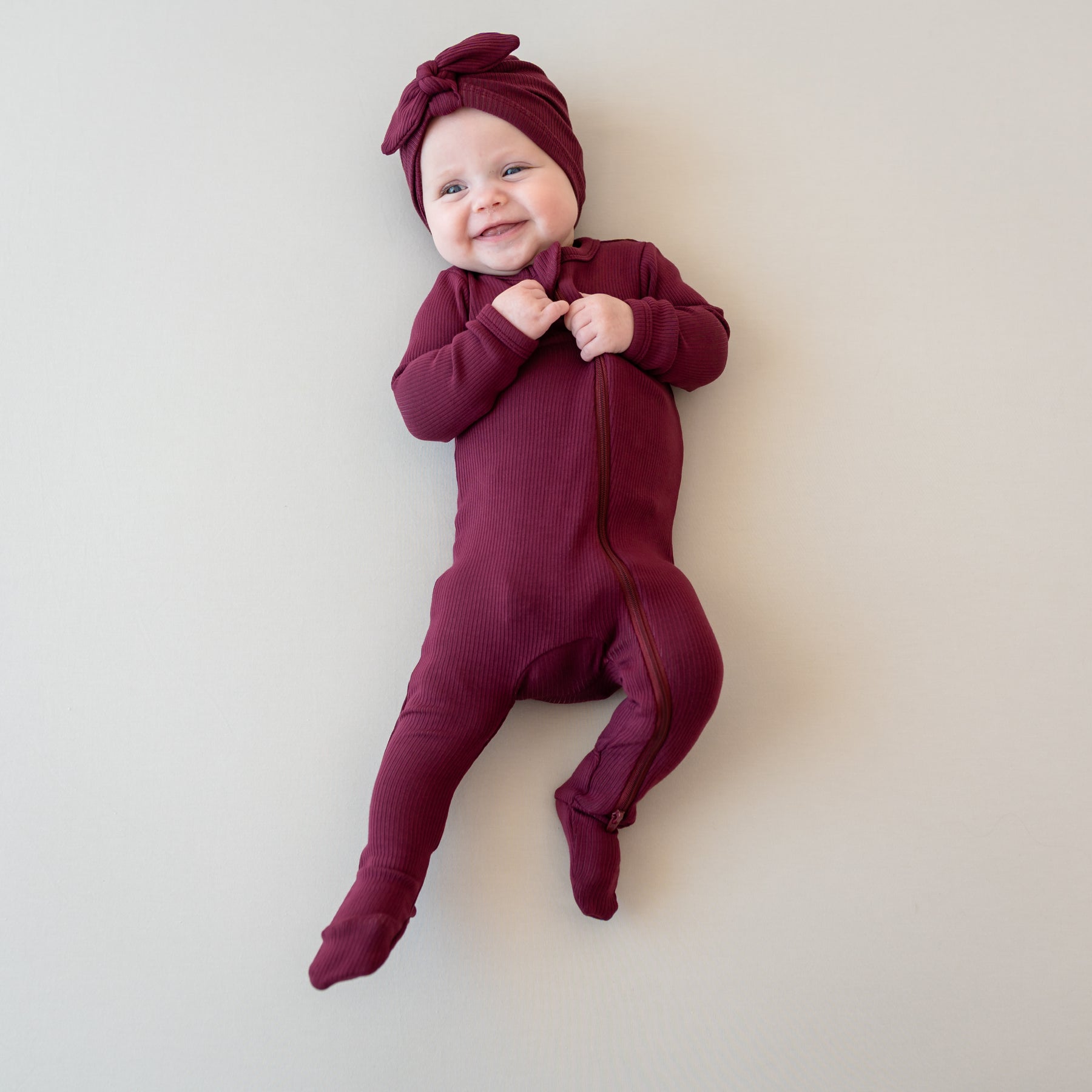 Smiling infant wearing the Ribbed Zipper Footie in Burgundy with matching ribbed headwrap laying on a neutral color backdrop