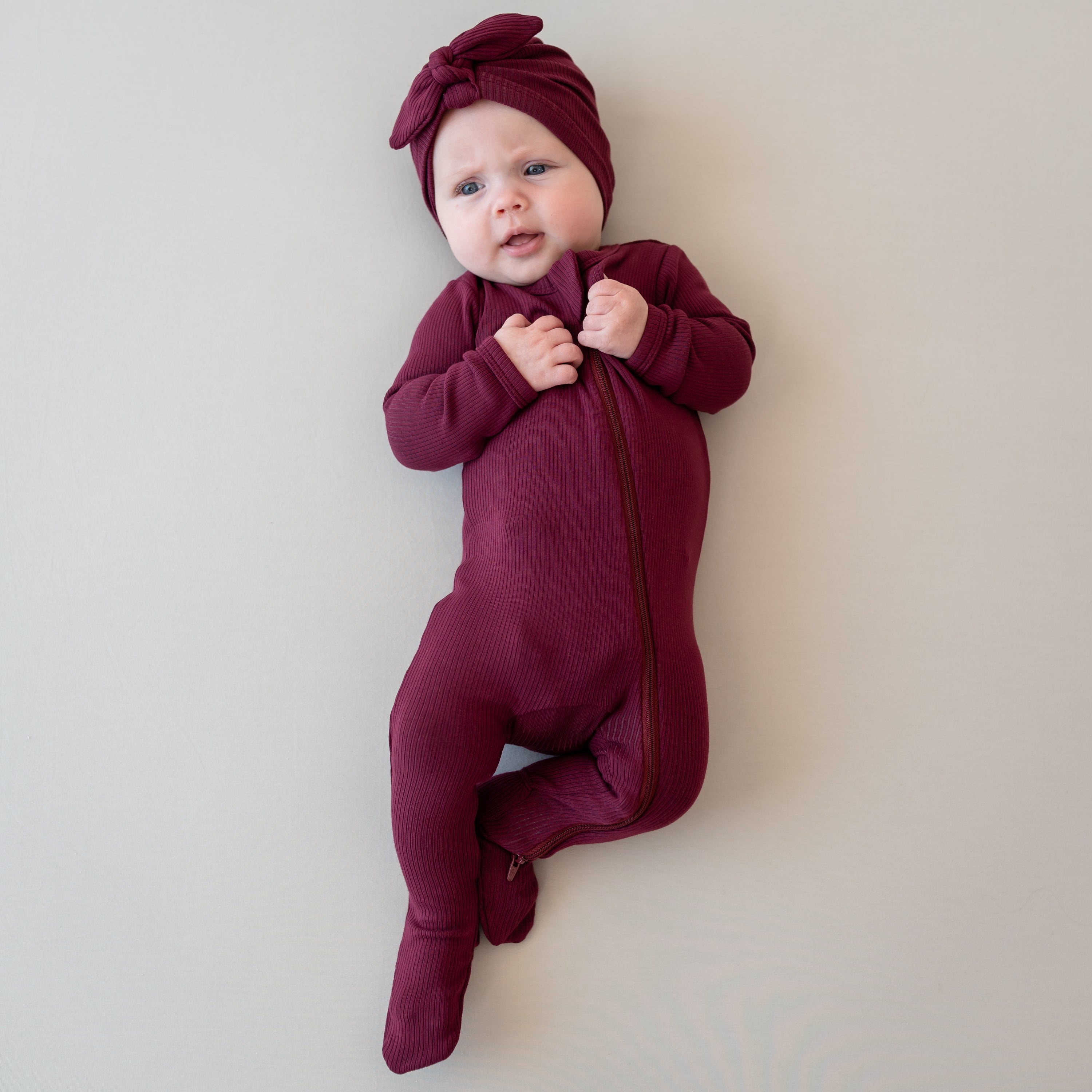 Infant laying down wearing the Ribbed Zipper Footie in Burgundy with matching ribbed headwrap on a light neutral backdrop
