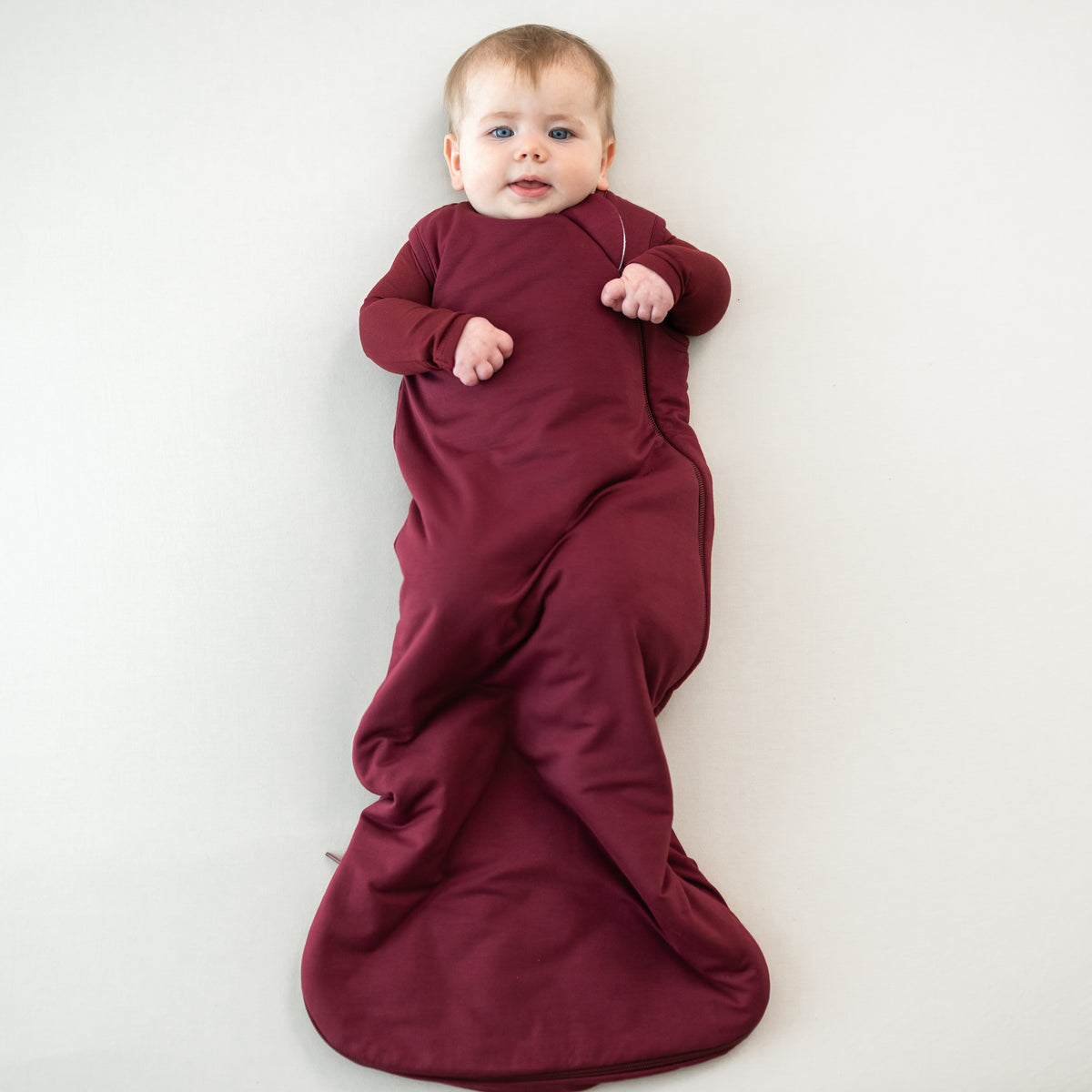 Infant lying down wearing the Sleep Bag in Burgundy 1.0 with matching long sleeve romper on underneath