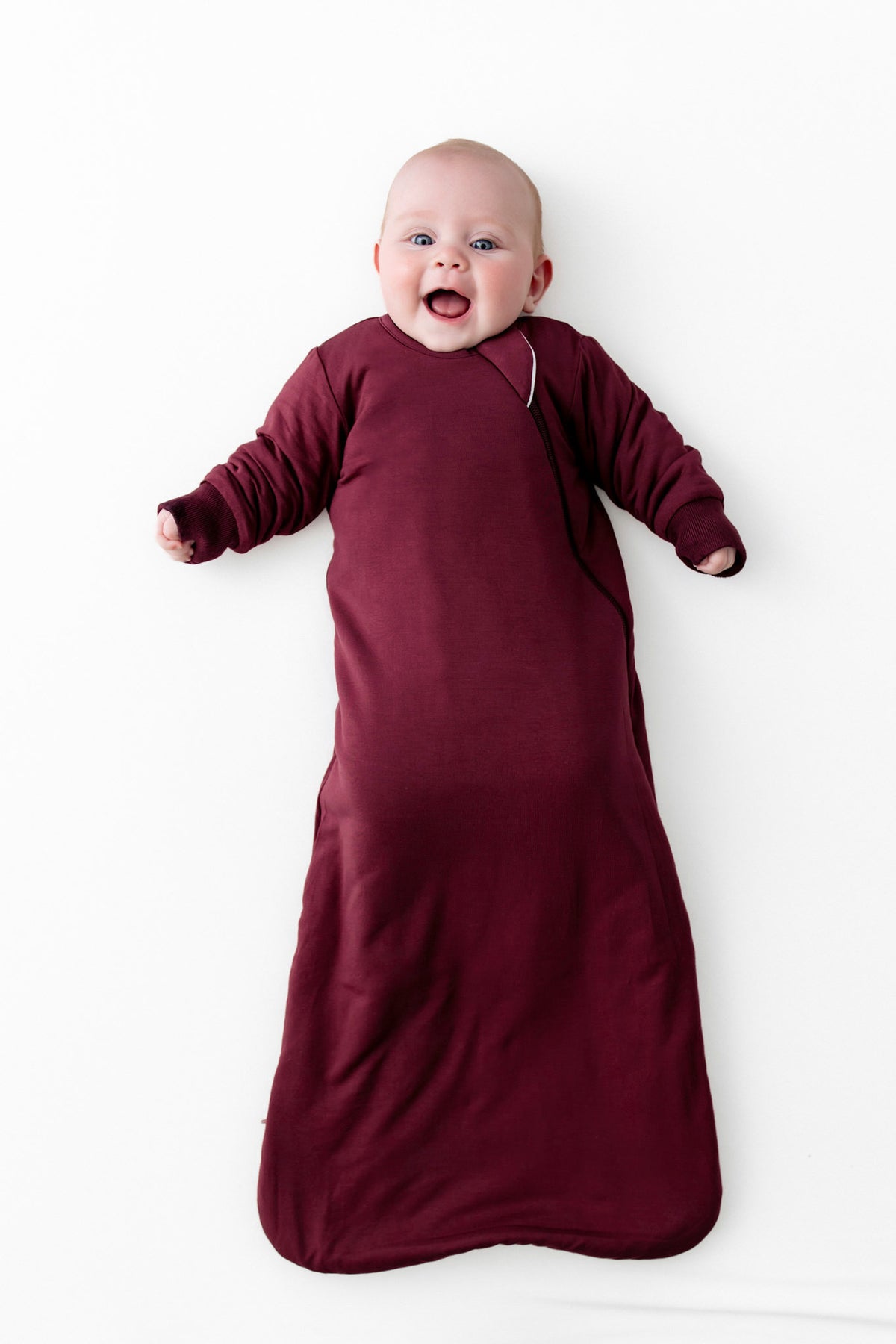 A baby lying on their back wearing a long-sleeve burgundy sleep sack against a plain white background.