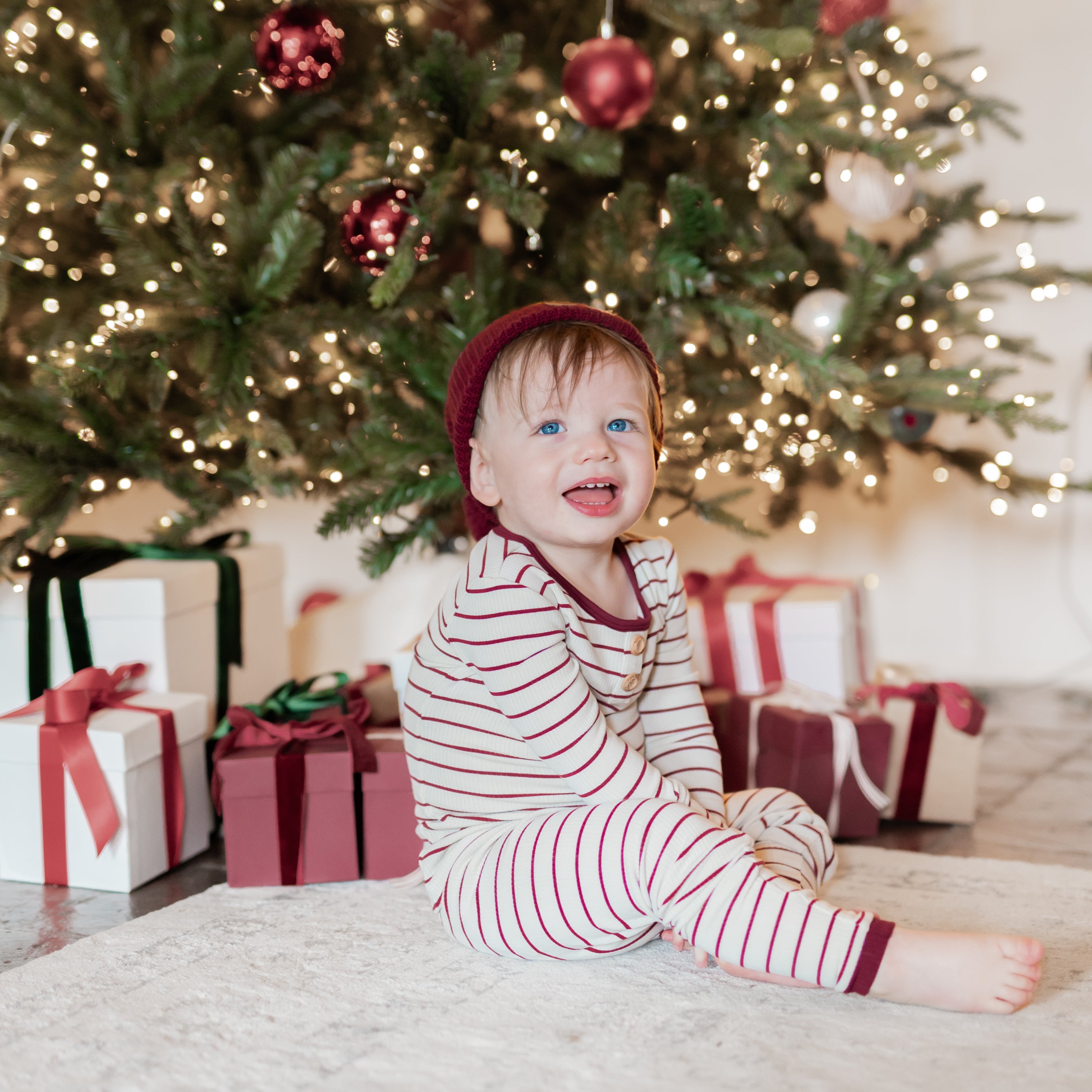 Toddler sitting on the floor wearing the Ribbed Henley Set in Burgundy Stripe in front of a decorated christmas tree with gifts underneath