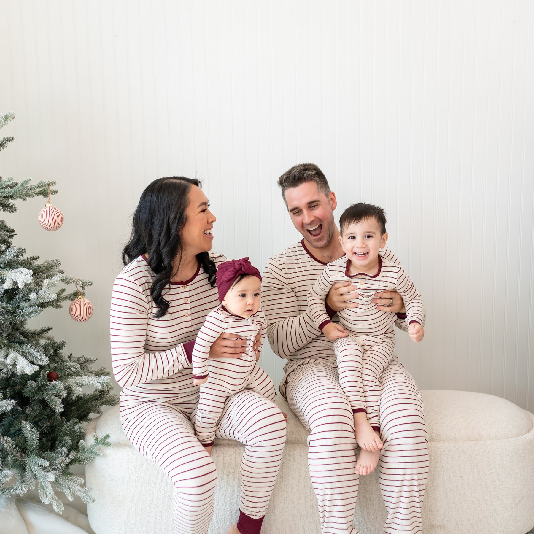 Family of four sitting on a cream ottoman beside a decorate Christmas tree wearing Ribbed Henley Sets in Burgundy Stripe