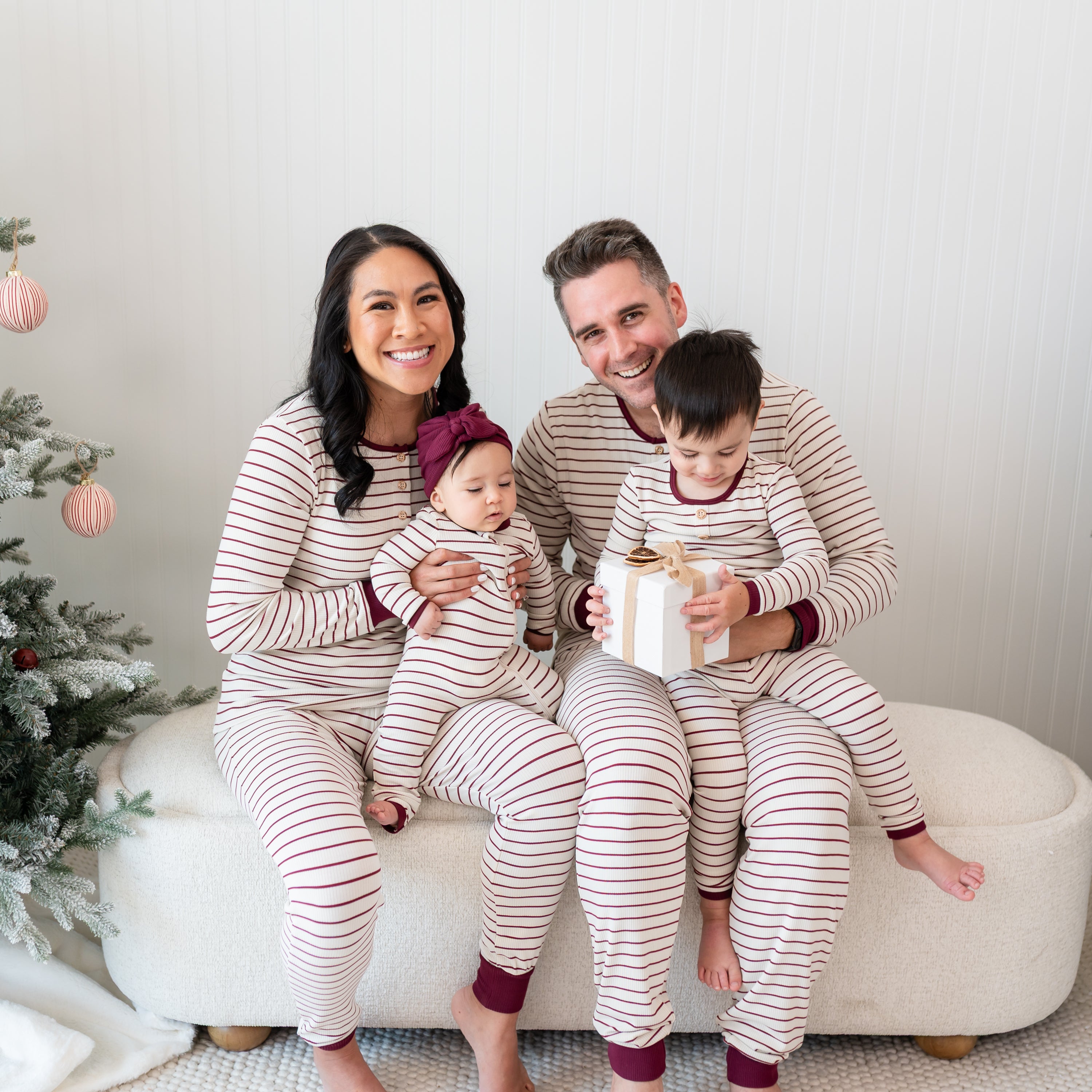 Family of four sitting on a cream ottoman wearing Ribbed Henley Sets in Burgundy Stripe