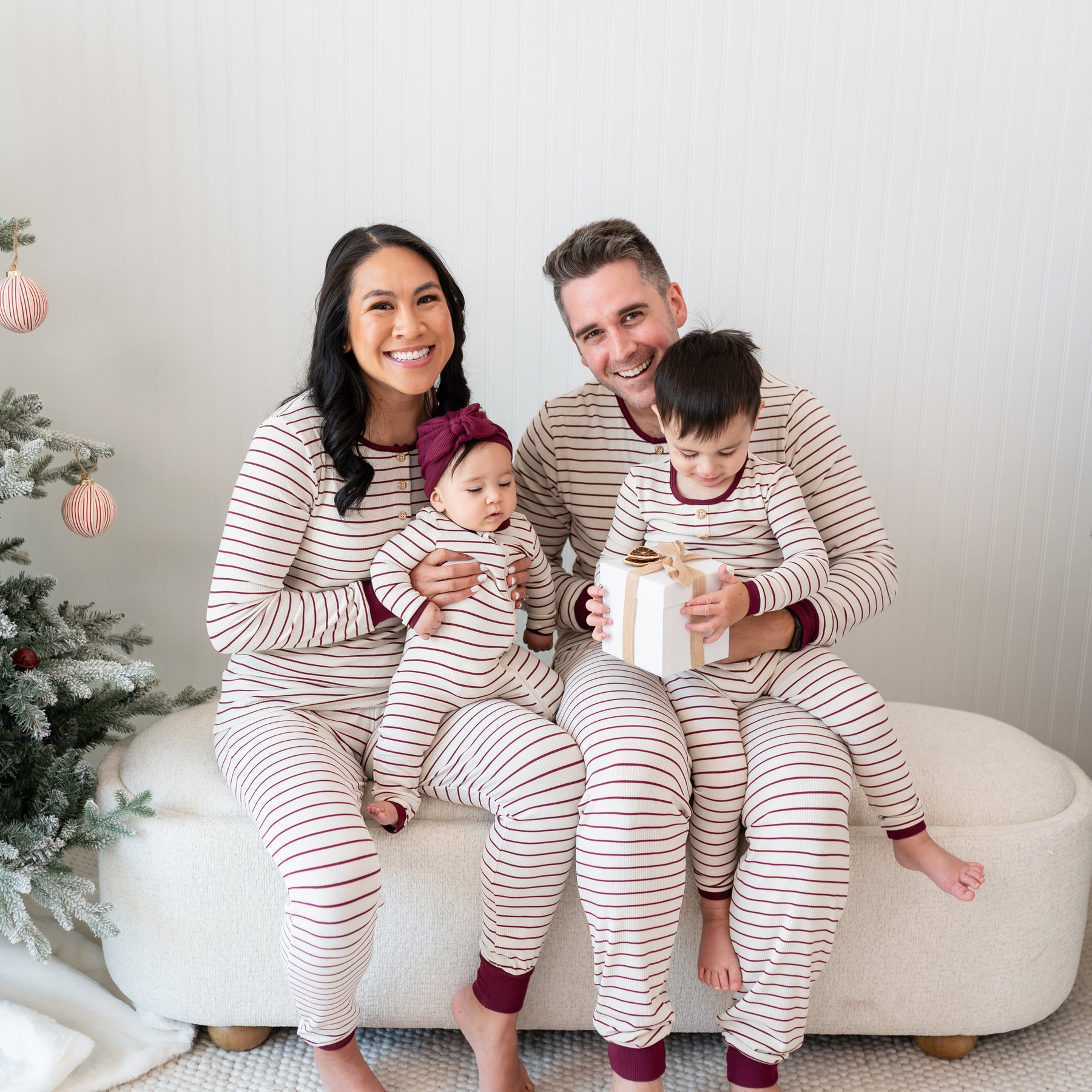 Family of four sitting on a cream ottoman wearing Ribbed Henley Sets in Burgundy Stripe