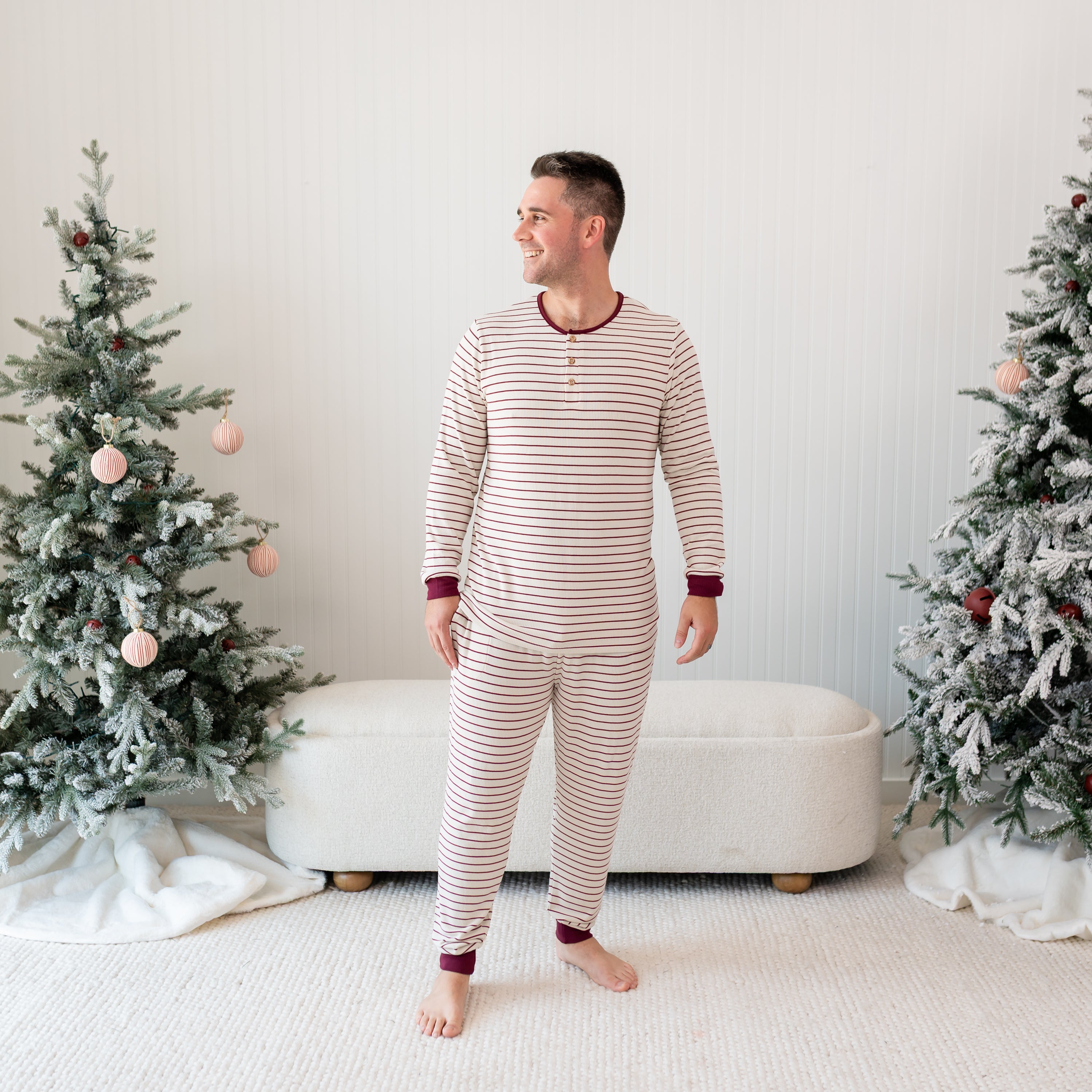 Male smiling looking off to the side standing in front of a cream ottoman and two decorated Christmas trees wearing the Men's Ribbed Henley Set in Burgundy Stripe