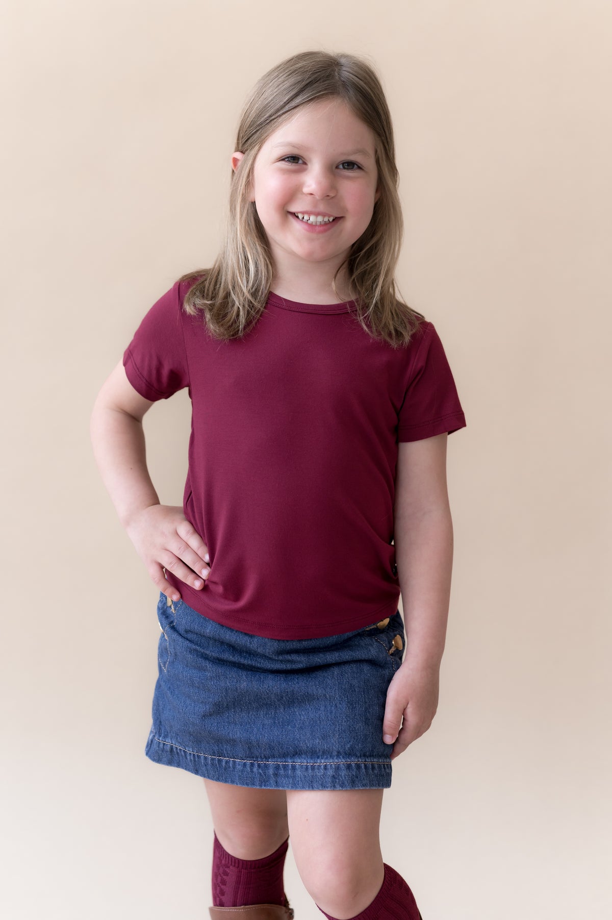 Girl wearing Toddler Basic Tee in Burgundy on blue skirt and burgundy knee high socks and boots on a beige background