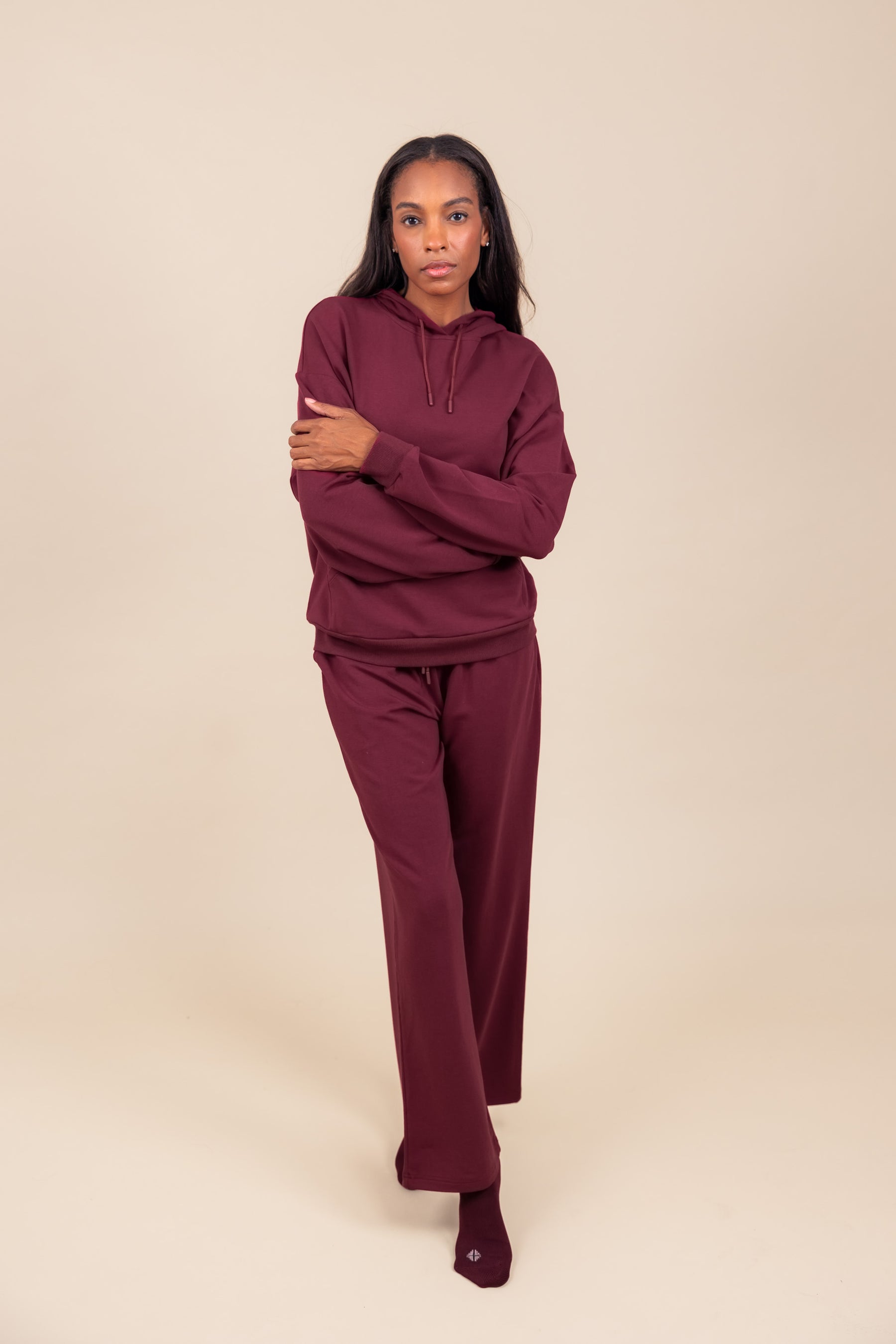 Woman wearing Women's Hoodie in Burgundy color with crossed hands over similar color women's sweatpants on a beige background