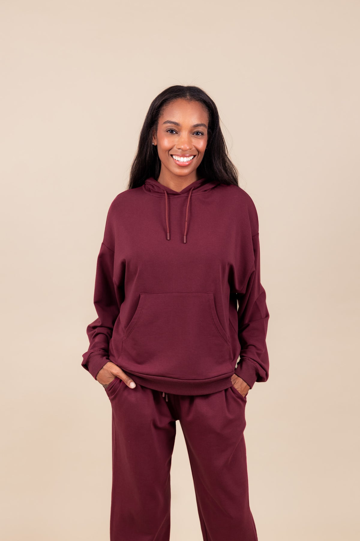 Woman smiling in Women's Hoodie in Burgundy color over similar color women's sweatpants with pockets on a beige background