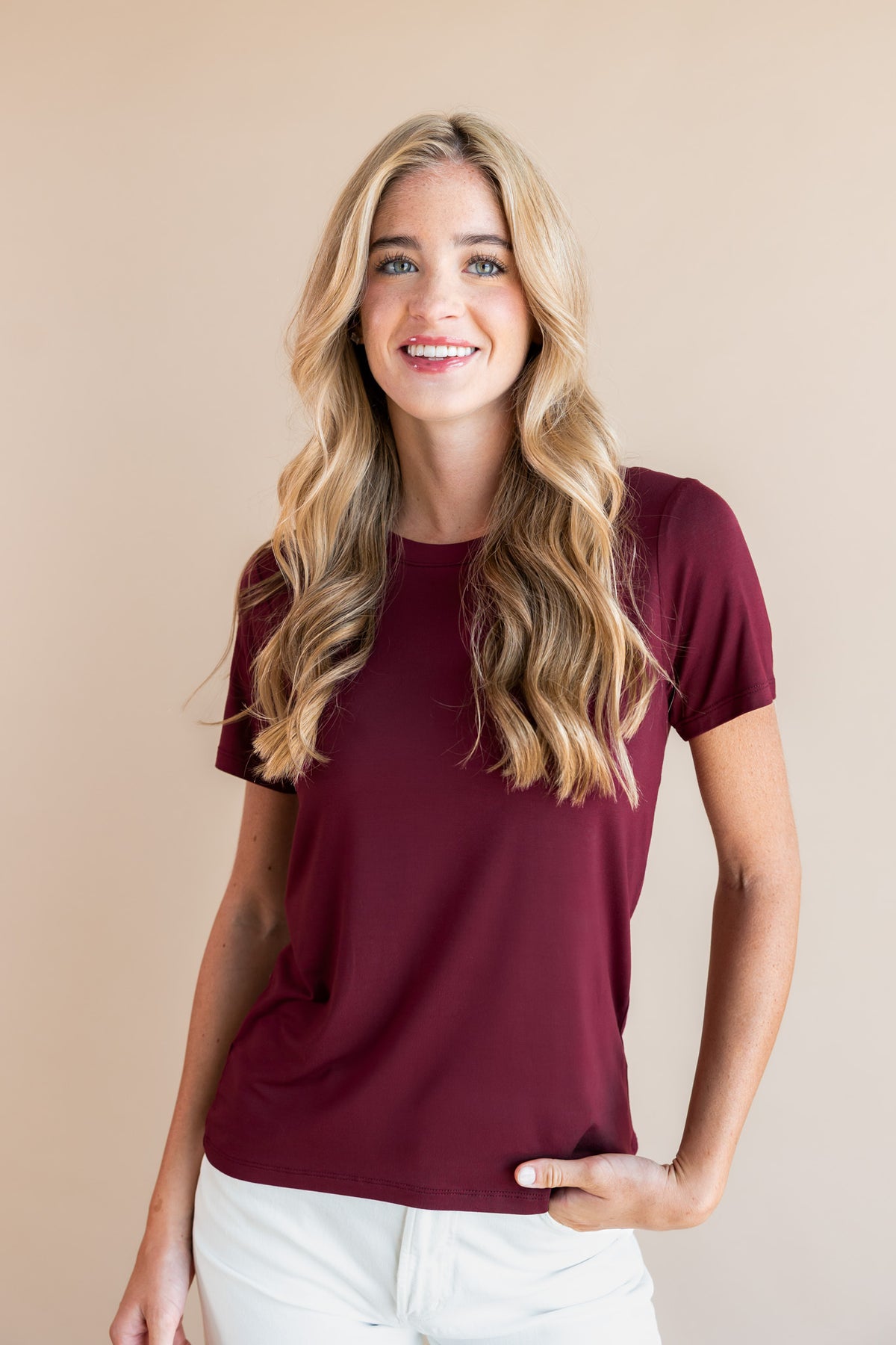 A smiling blonde woman wearing a short-sleeve crewneck t-shirt in burgundy and white pants, standing against a solid beige studio background.