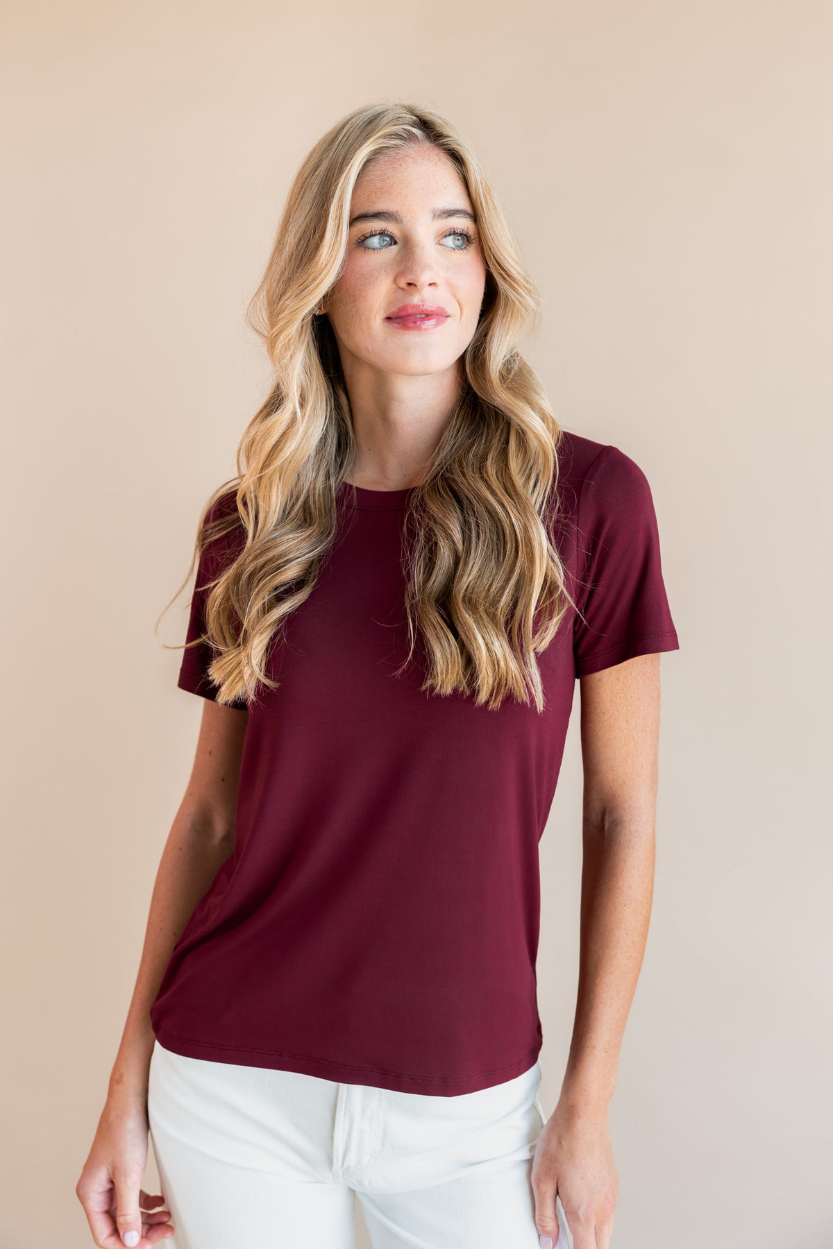 A woman with long wavy blonde hair wearing a short-sleeve crewneck t-shirt in burgundy and white pants, standing against a solid beige studio background.