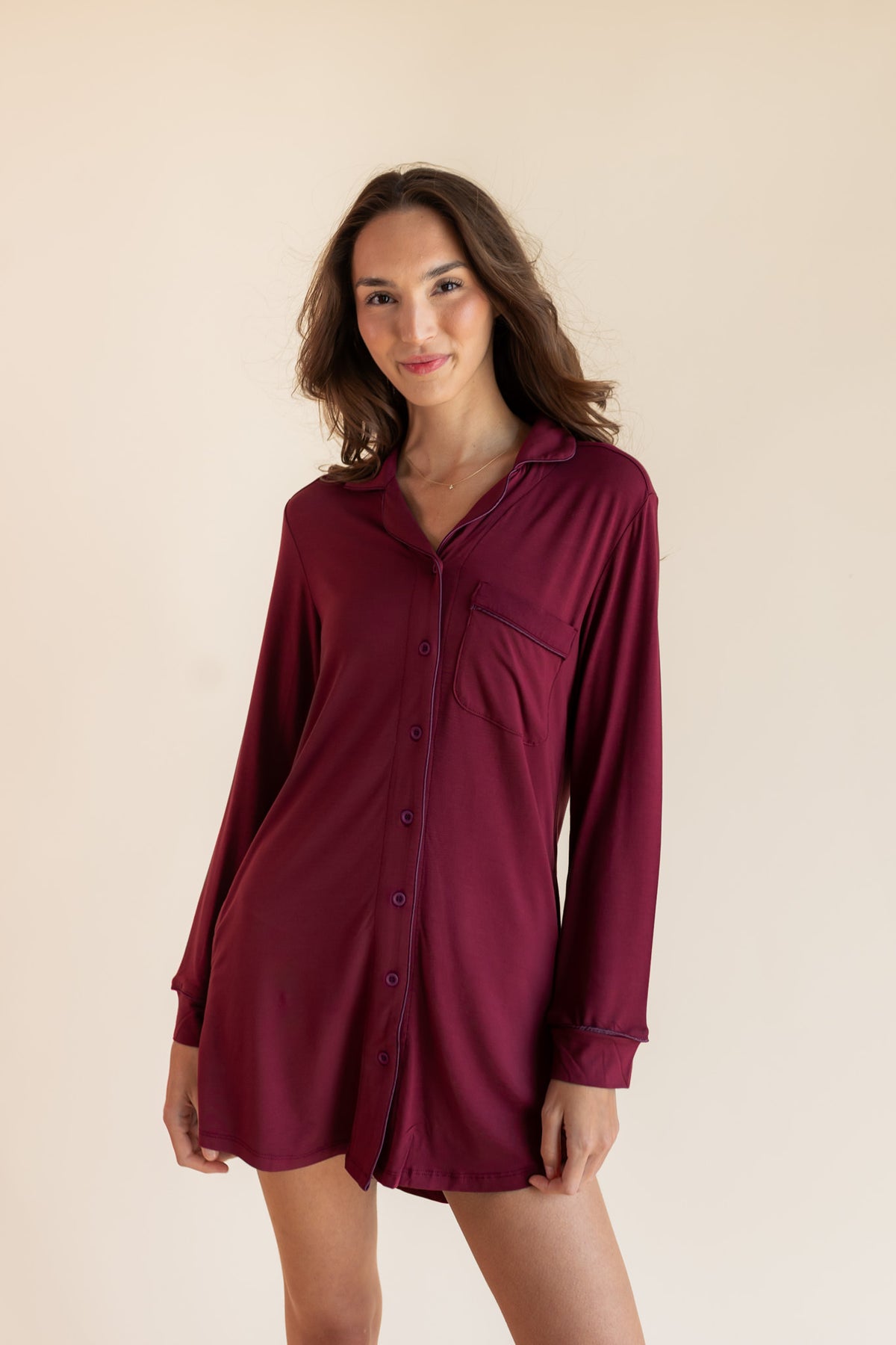 A woman modeling a long-sleeve button-front shirt dress in deep burgundy against a plain beige studio background.