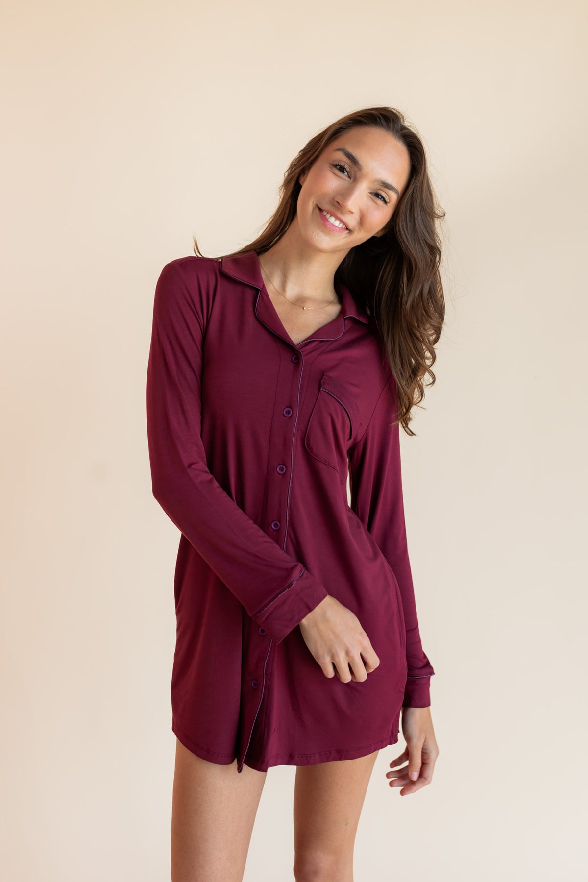 A woman modeling a long-sleeve burgundy button-up shirt dress against a solid beige studio background.