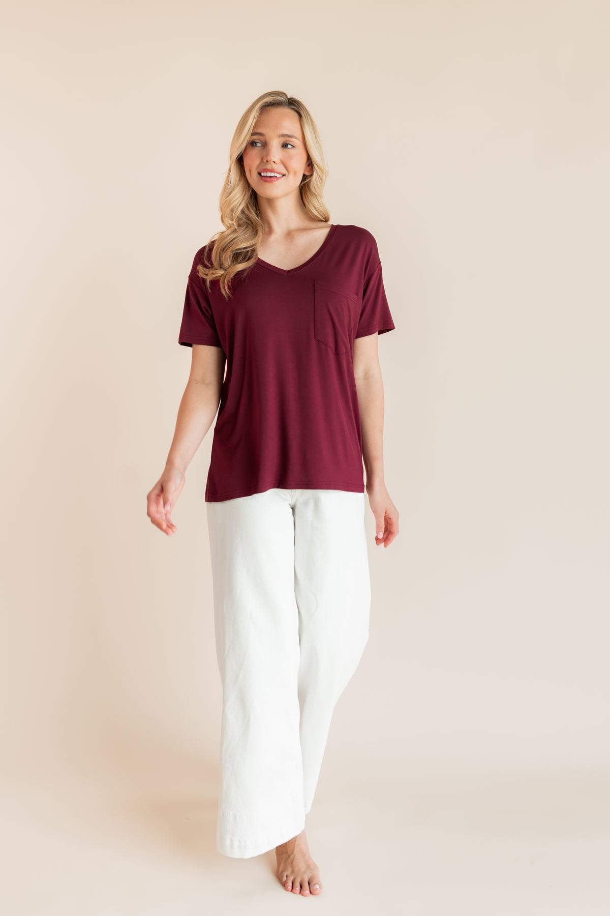 Model posing in Women's Oversized V-Neck Tee in Burgundy over white pants on a beige background