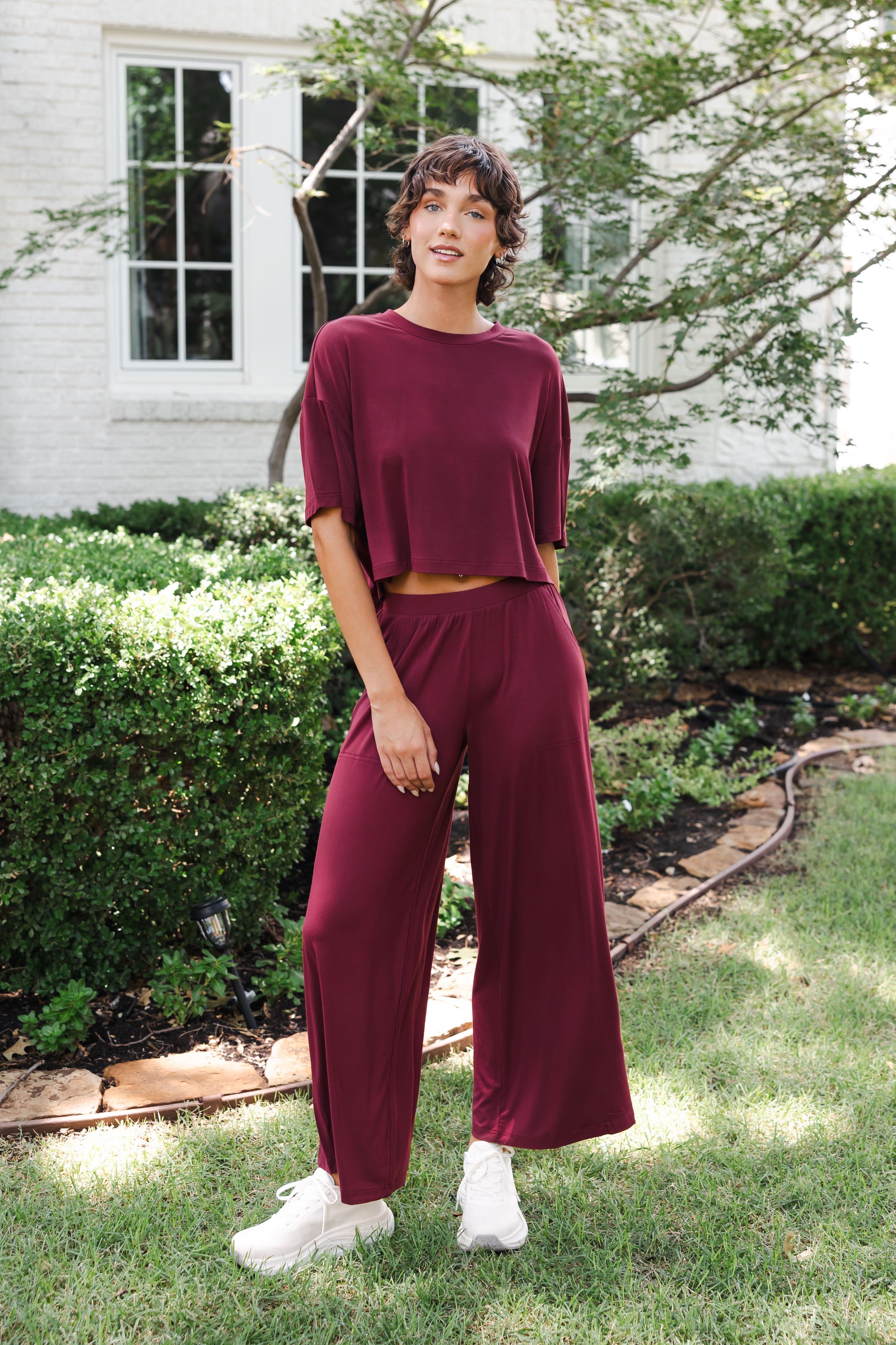 Female model standing in garden wearing the Women's Wide Leg Pant in Burgundy and matching short sleeve tee on white shoes