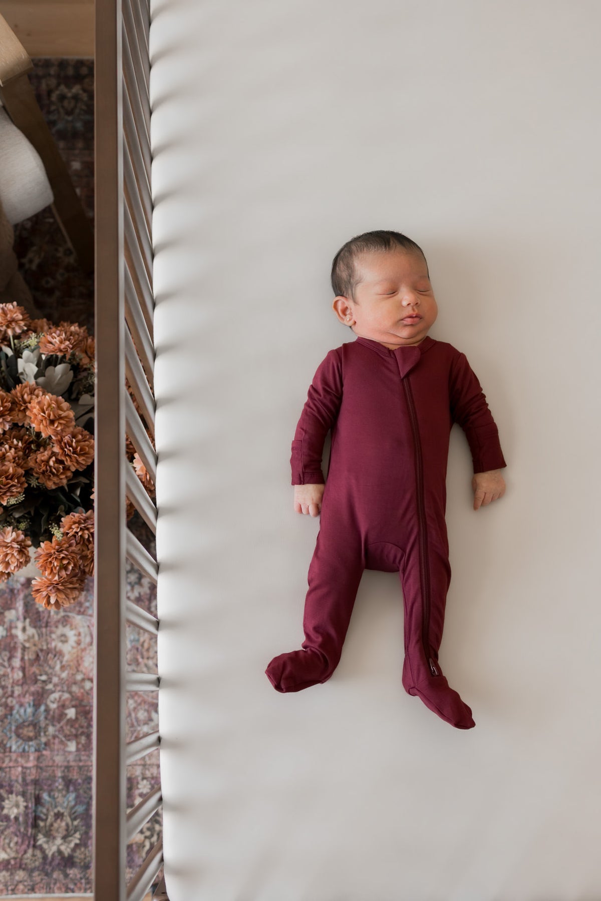 A sleeping newborn baby wearing a burgundy footed sleeper, lying on a white mattress inside a crib.