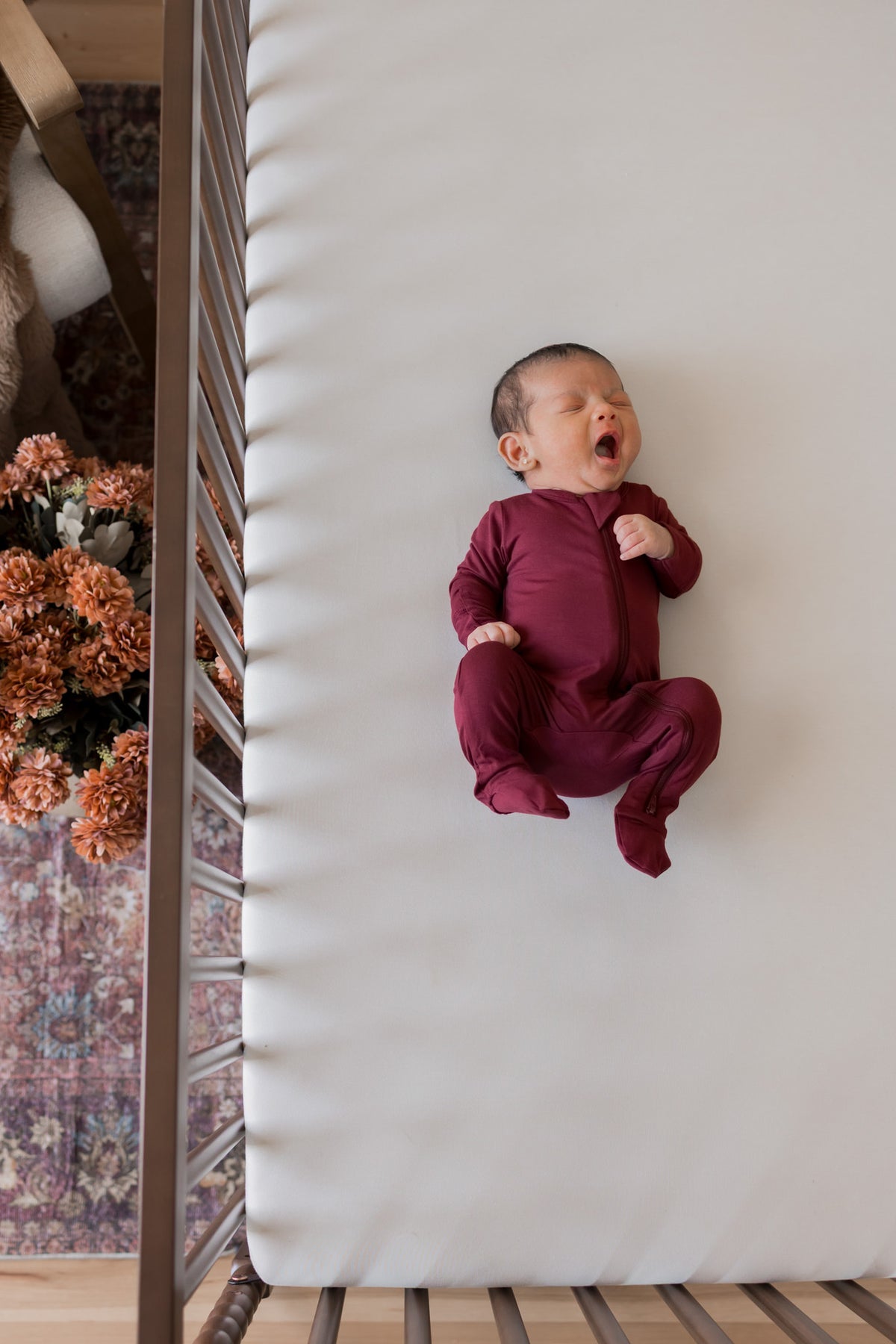 A newborn baby wearing a burgundy long-sleeve footed sleeper, lying on their back on a white mattress inside a wooden crib.