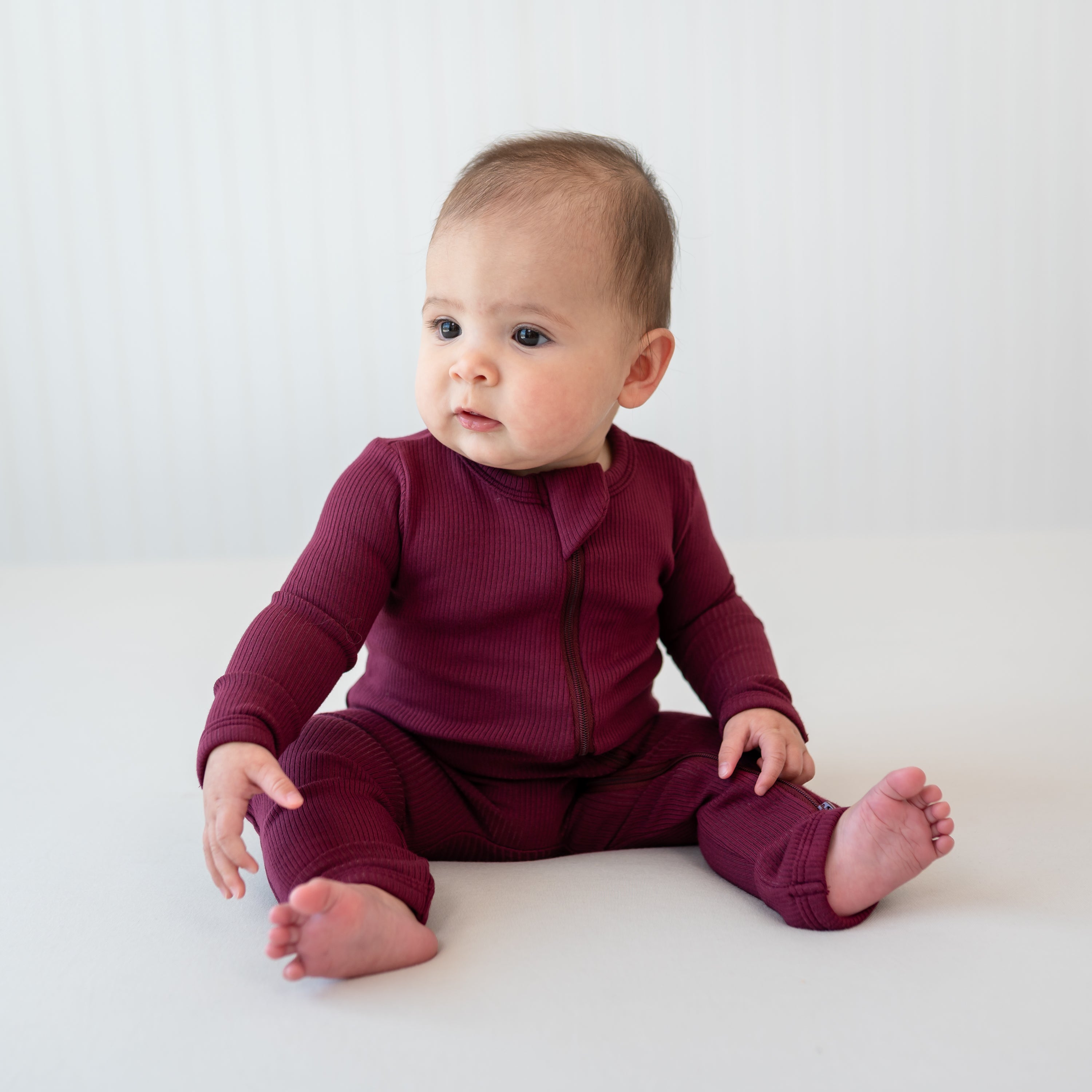 Infant sitting wearing the Ribbed Zipper Romper in Burgundy in front of a light colored backdrop