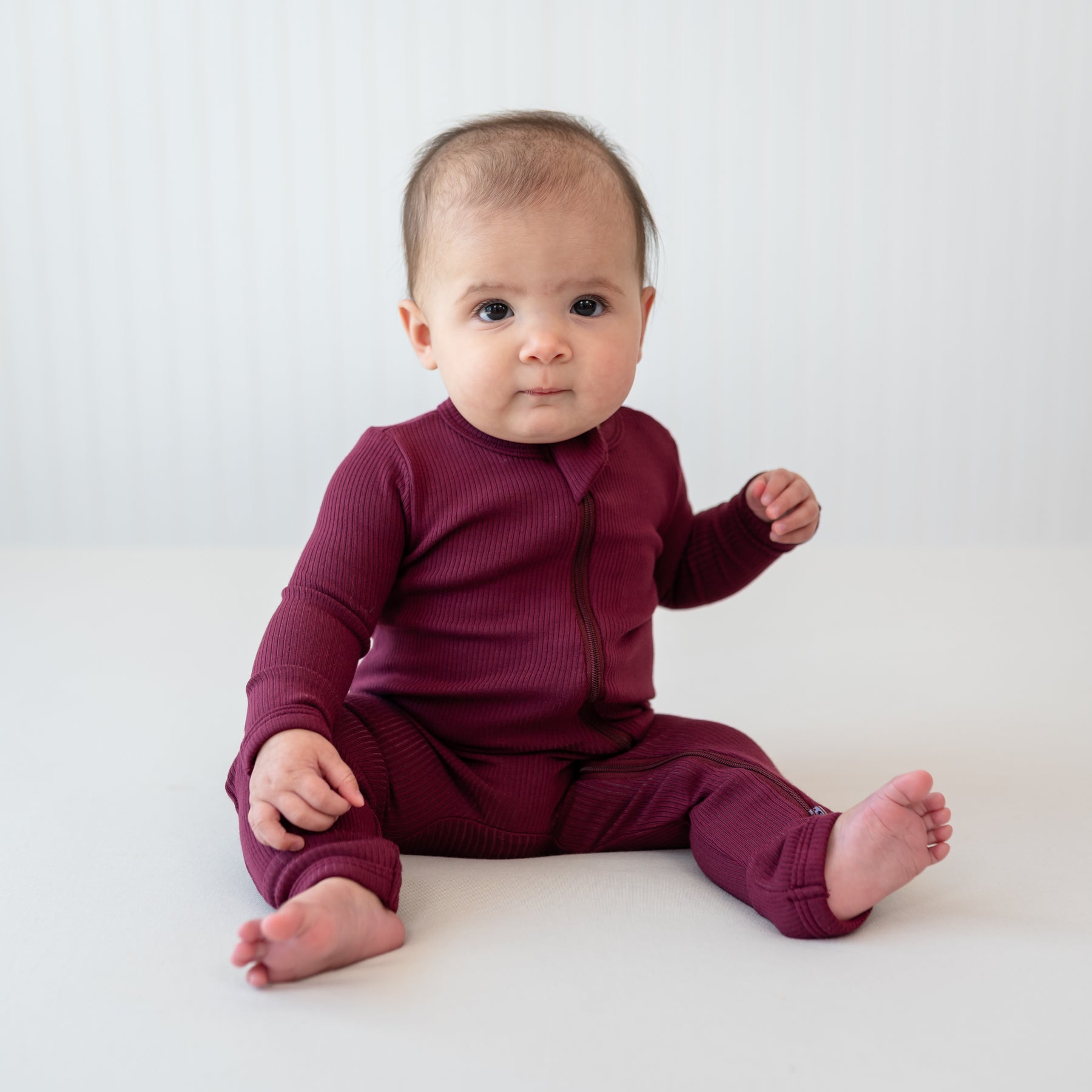 Infant sitting wearing the Ribbed Zipper Romper in Burgundy in front of a light colored backdrop