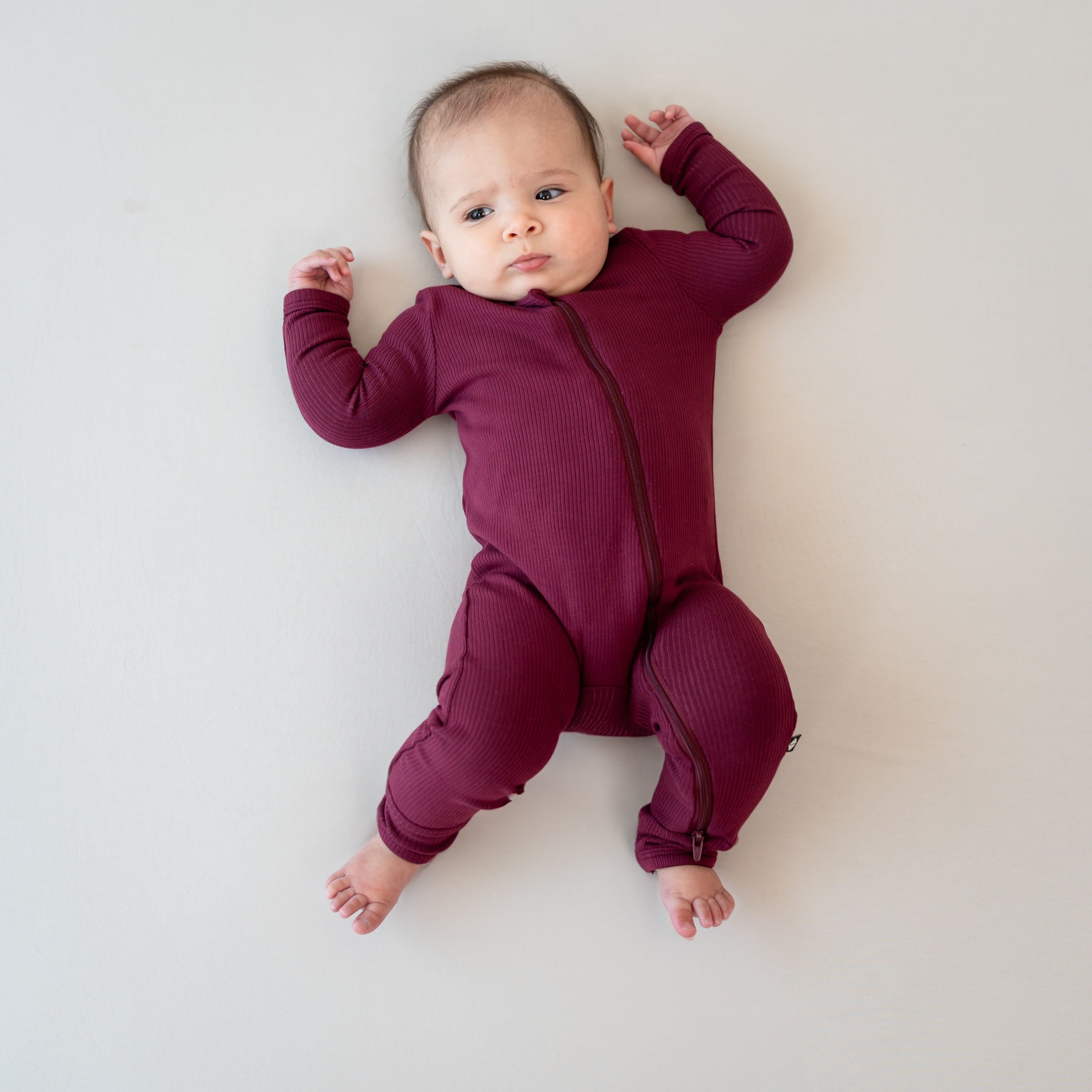 Infant laying on a neutral light colored background wearing the Ribbed Zipper Romper in Burgundy