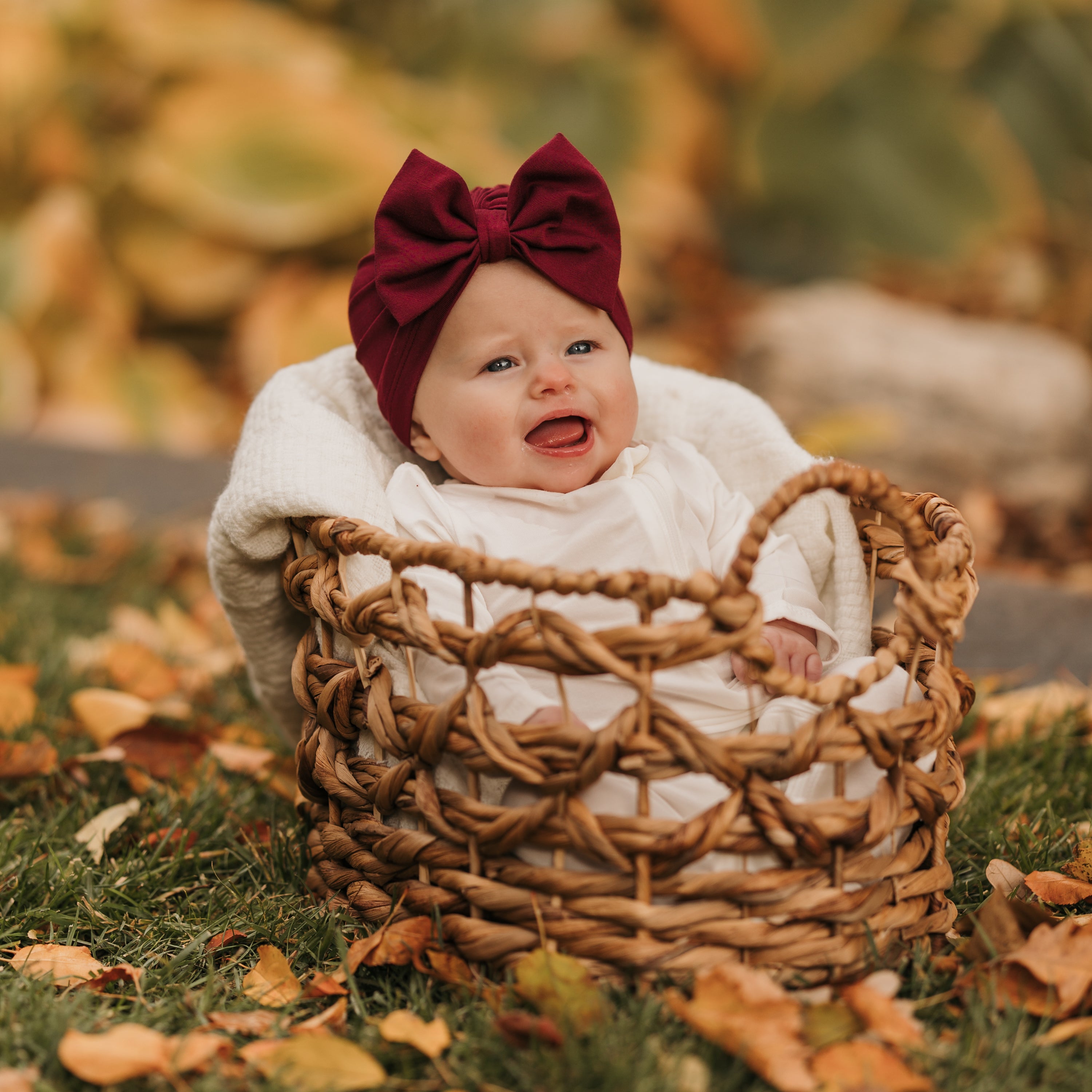 Smiling infant sitting in a wicker basket in grass and leaves wearing the Bow Headwrap in Burgundy and white romper