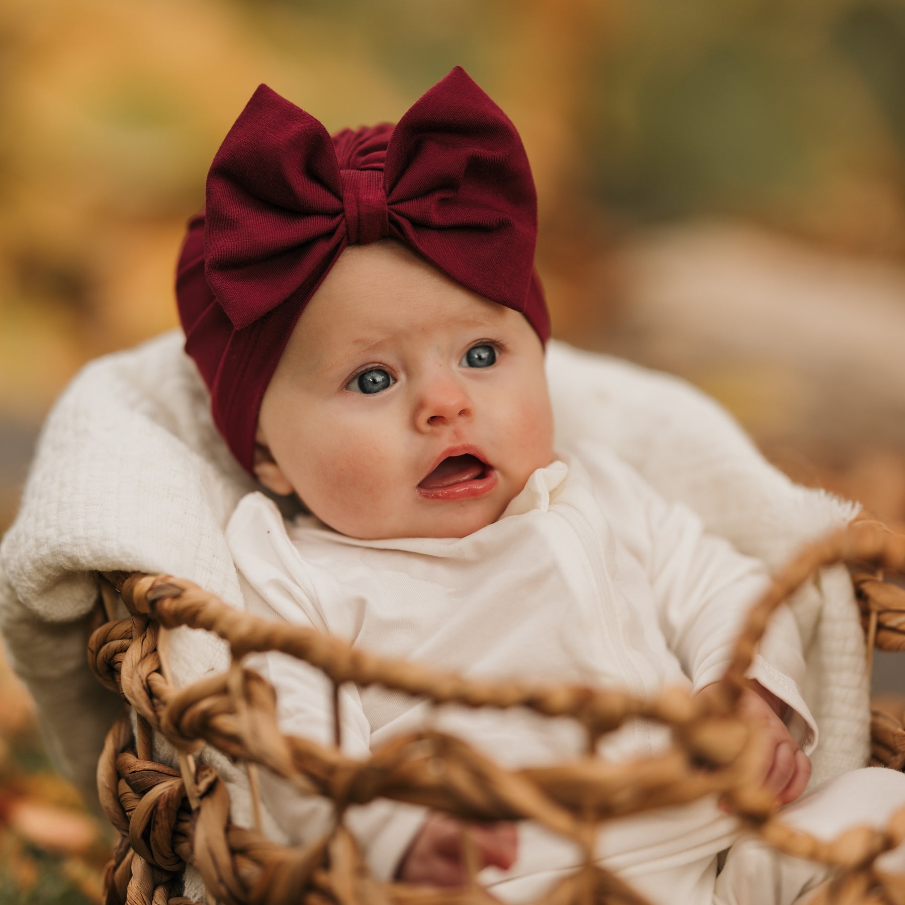 Infant sitting in a wicker basket wearing the Bow Headwrap in Burgundy and a white romper